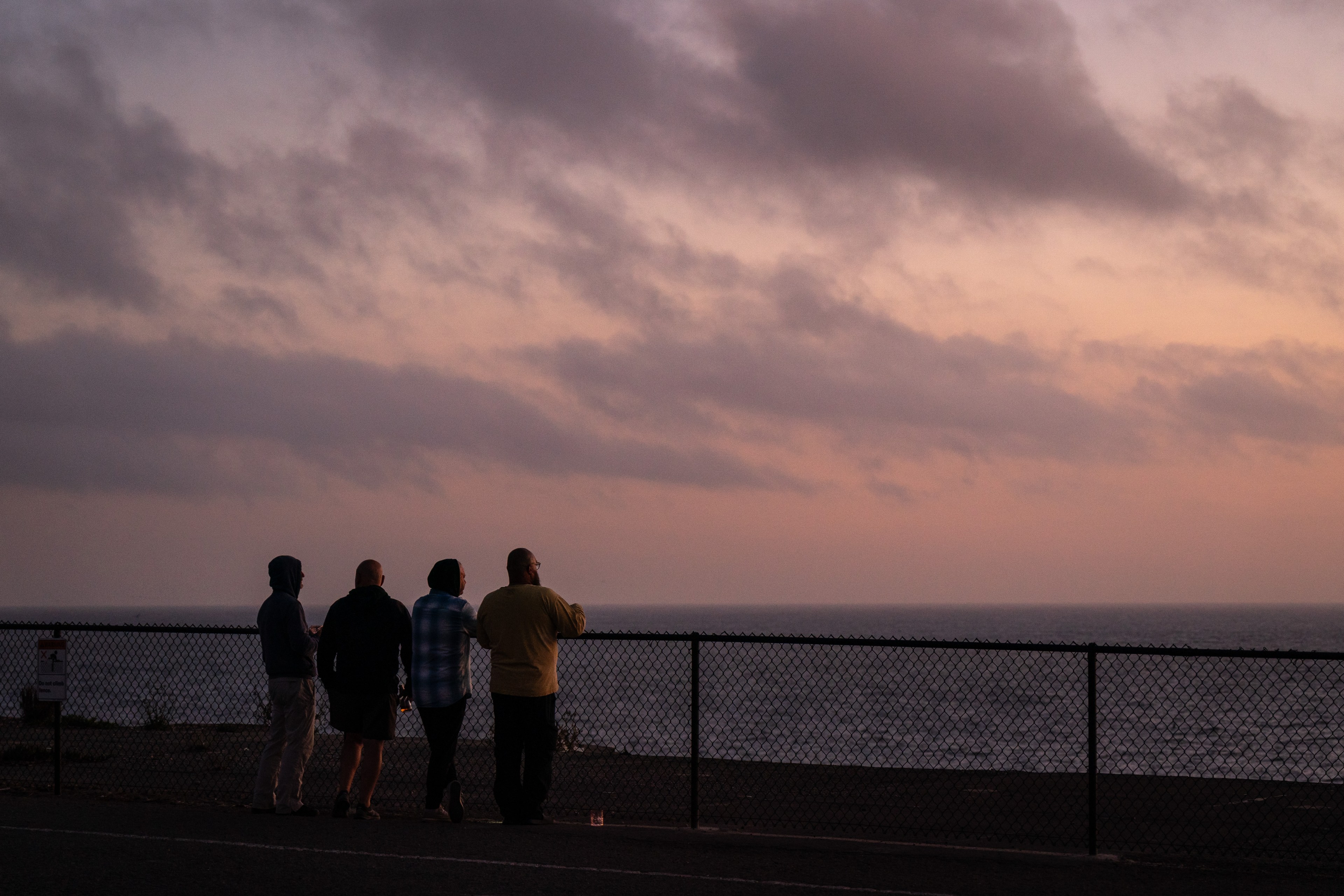 Four people stand beside a fence, gazing at an ocean during sunset under a cloudy sky, their silhouettes highlighted against the dusky backdrop.