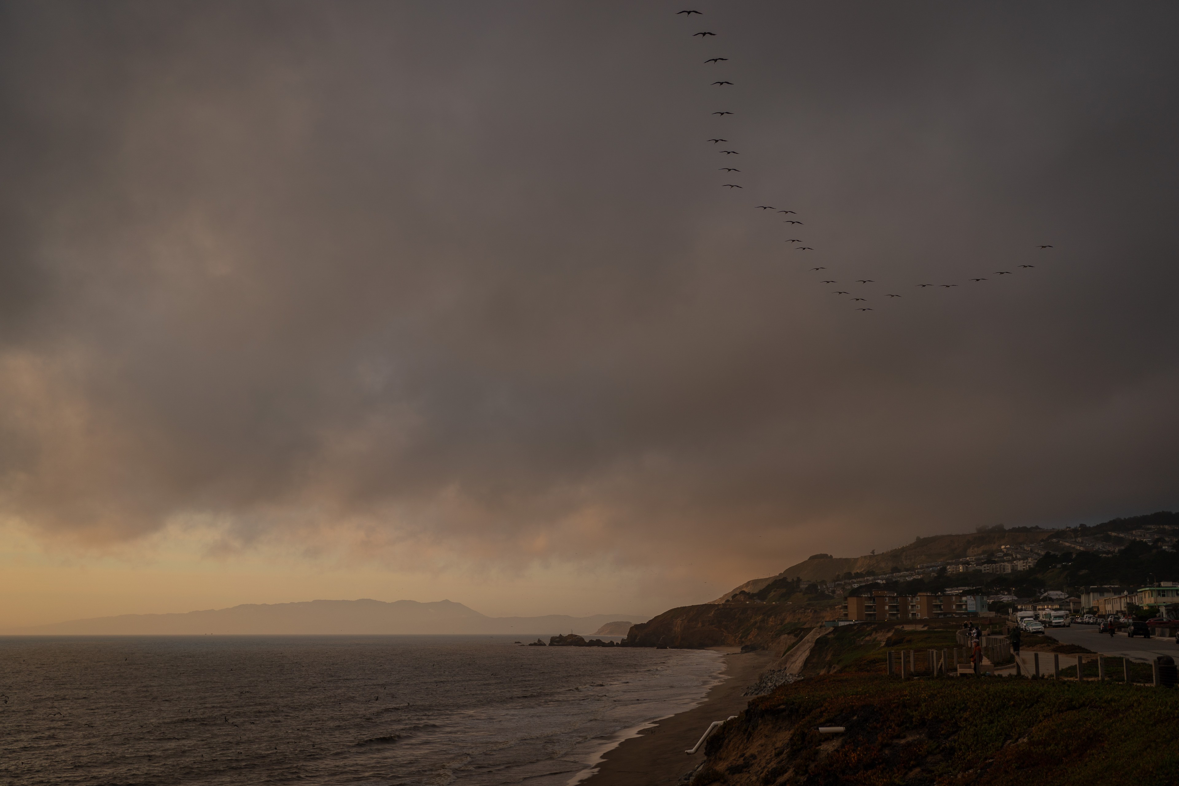 A coastal scene at dusk with a dark, cloudy sky, distant mountains, waves hitting the shore, and birds flying in a V formation above a beachside town.