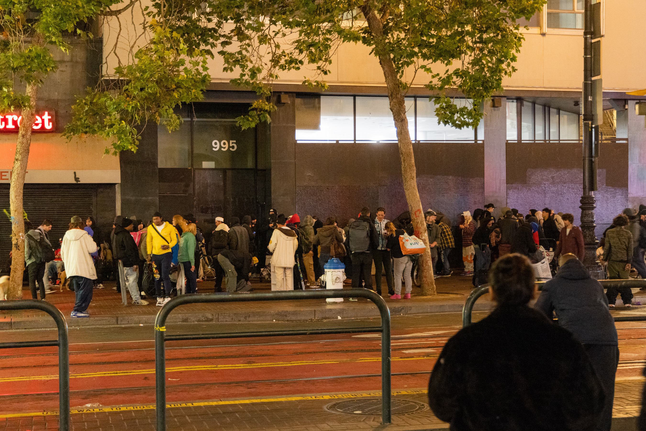 A large crowd of people is gathered on a sidewalk at night near a building with the number 995 above the entrance. Many are wearing coats and hats, and a few trees line the street.