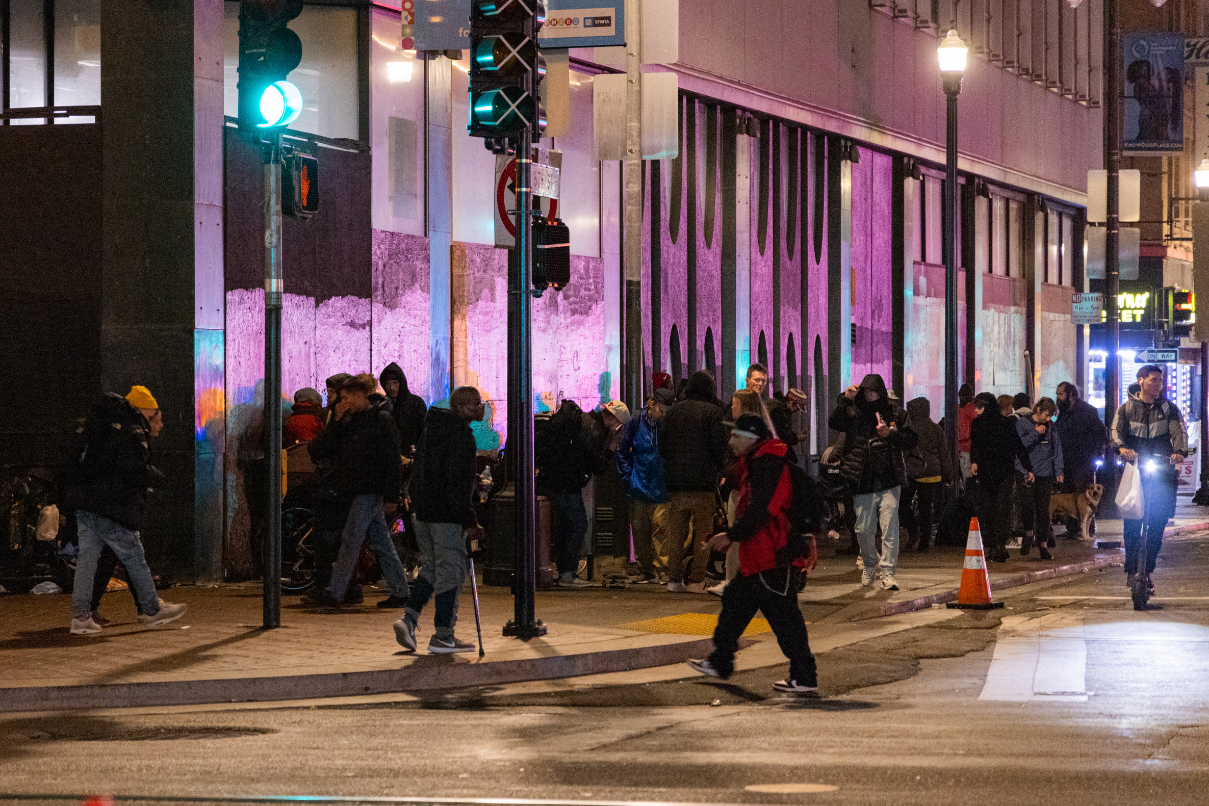 A crowded city sidewalk at night with people bundled up in jackets, a green traffic light, and a lit-up storefront casting purple light on a building wall.