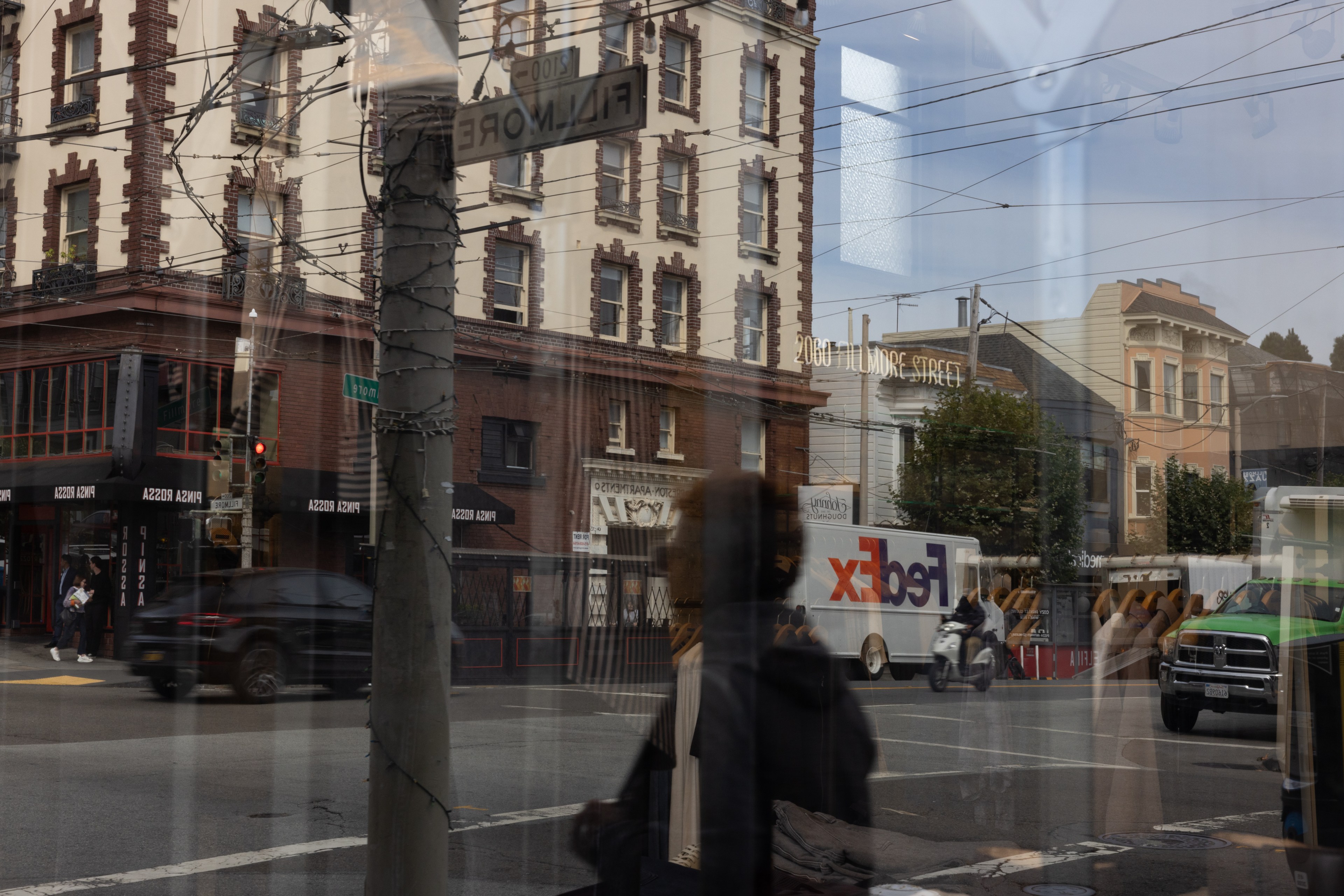 The image shows a busy urban street corner with buildings, a FedEx truck, a cyclist, and pedestrians. Reflections in the glass capture additional street scenes.