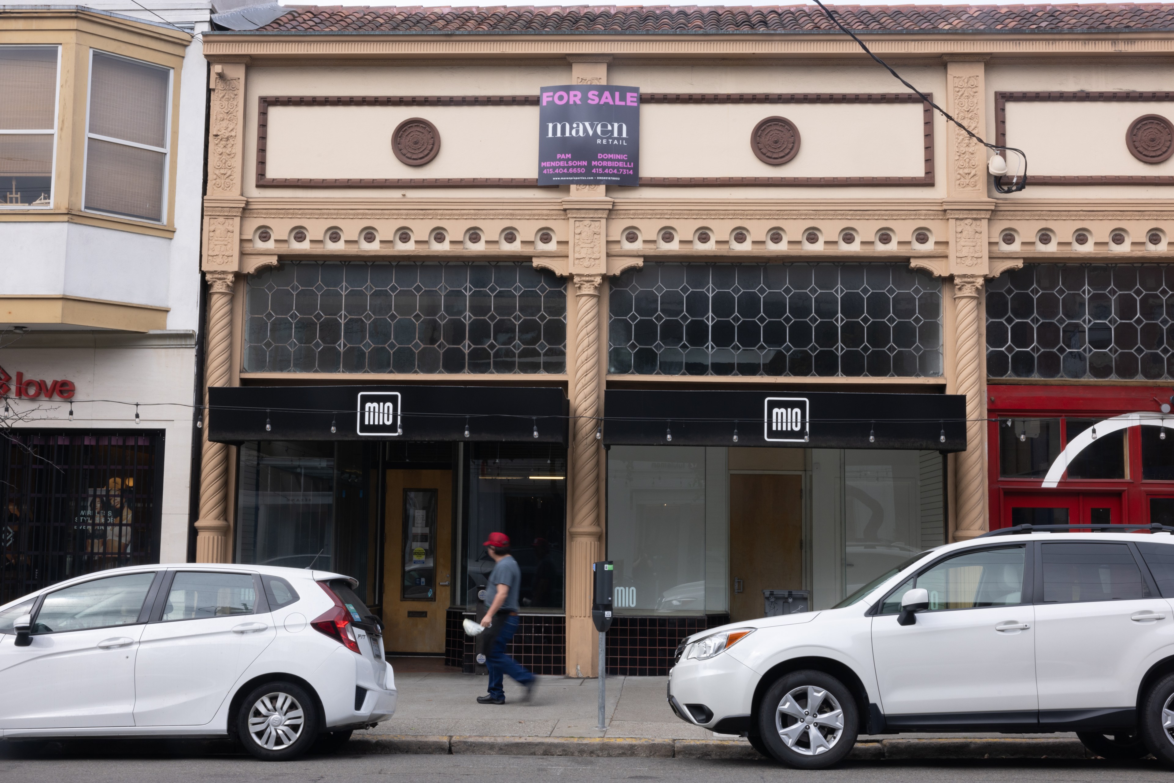 The image shows a street view of a storefront with a &quot;For Sale&quot; sign for Maven Retail. Two cars are parked in front, and a person is walking on the sidewalk.