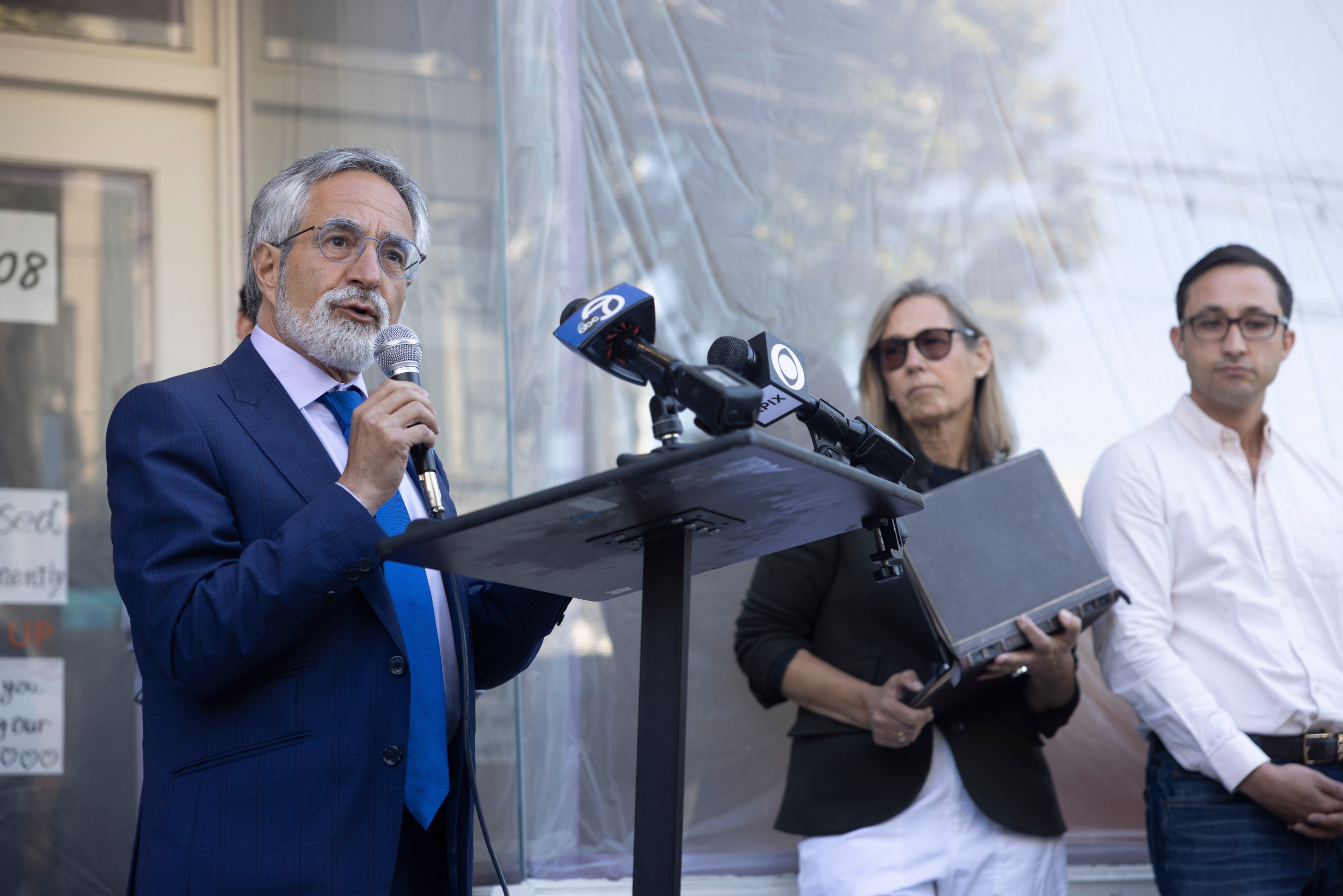 A man in a blue suit speaks into a microphone at a podium with cameras. Two people stand behind him; one holds a laptop, and the other has a neutral expression.