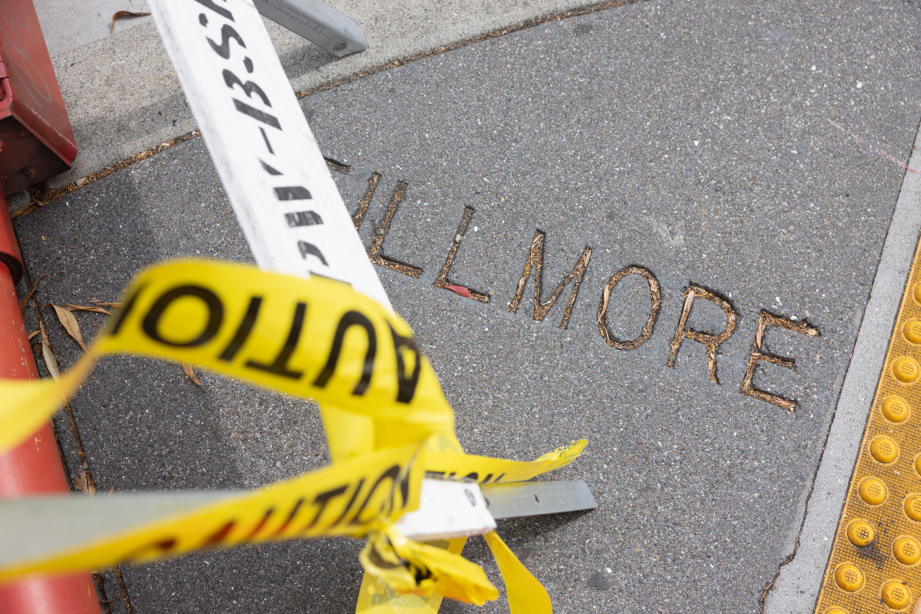 The image shows a concrete sidewalk with the word &quot;FILLMORE&quot; etched into it, partially covered by yellow caution tape and a metal barricade.