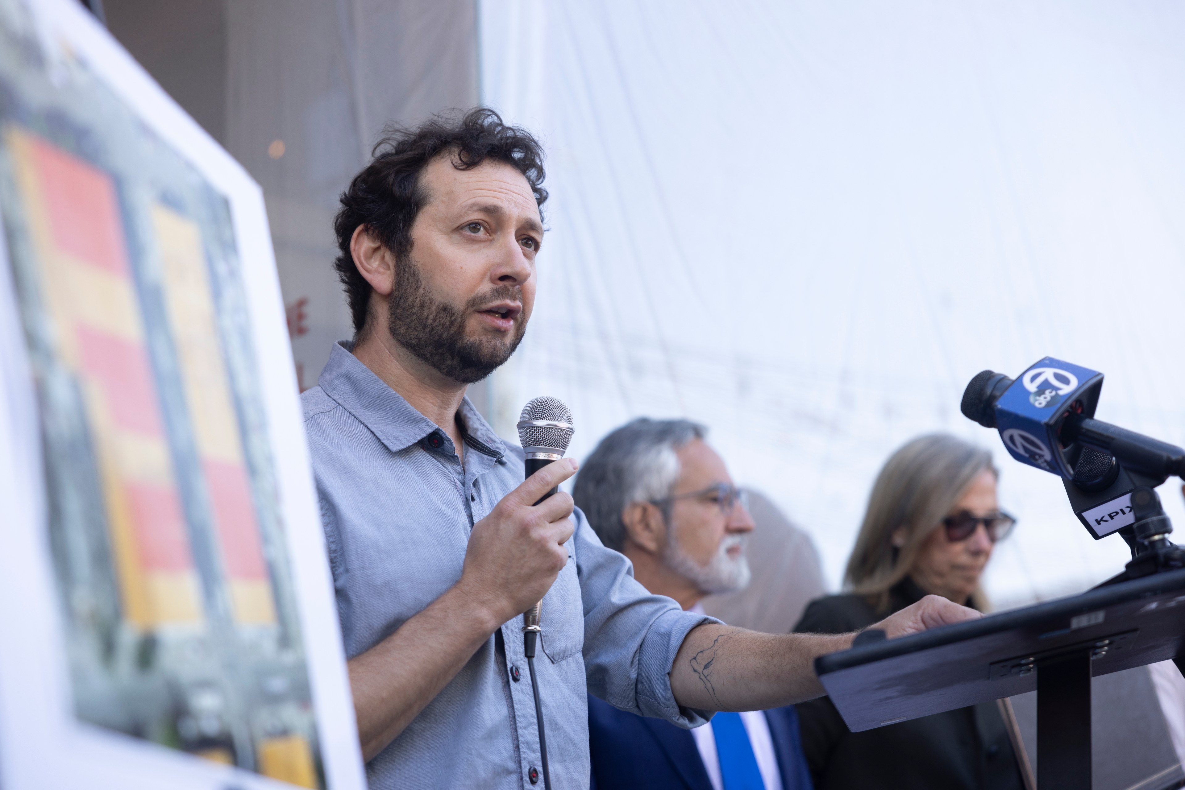 A man with a beard, holding a microphone, speaks at a podium. Two other individuals stand nearby, one with gray hair and the other wearing sunglasses.