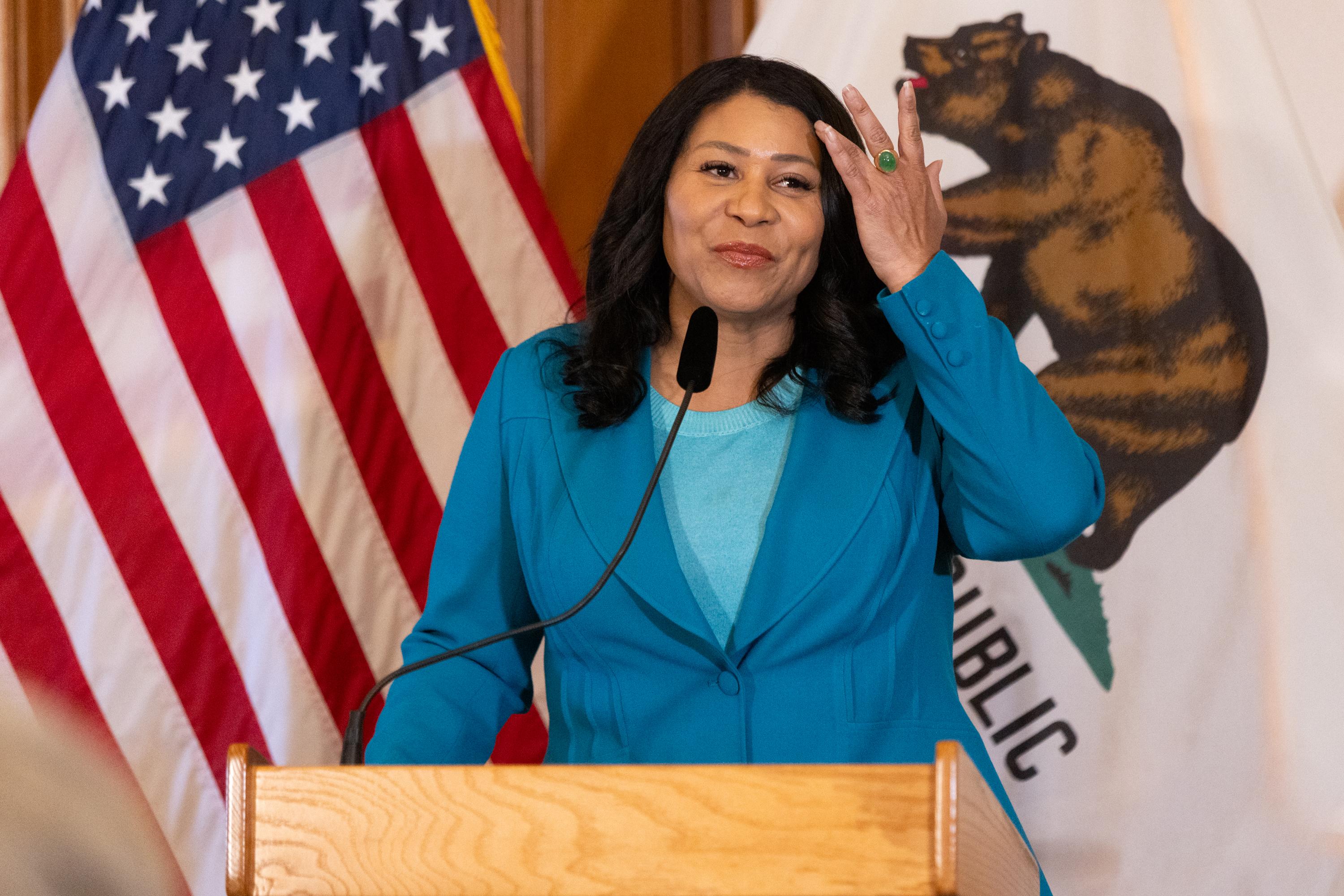 A woman in a teal outfit is speaking at a podium. Behind her are the American flag and the California state flag featuring a bear. She gestures with her right hand.