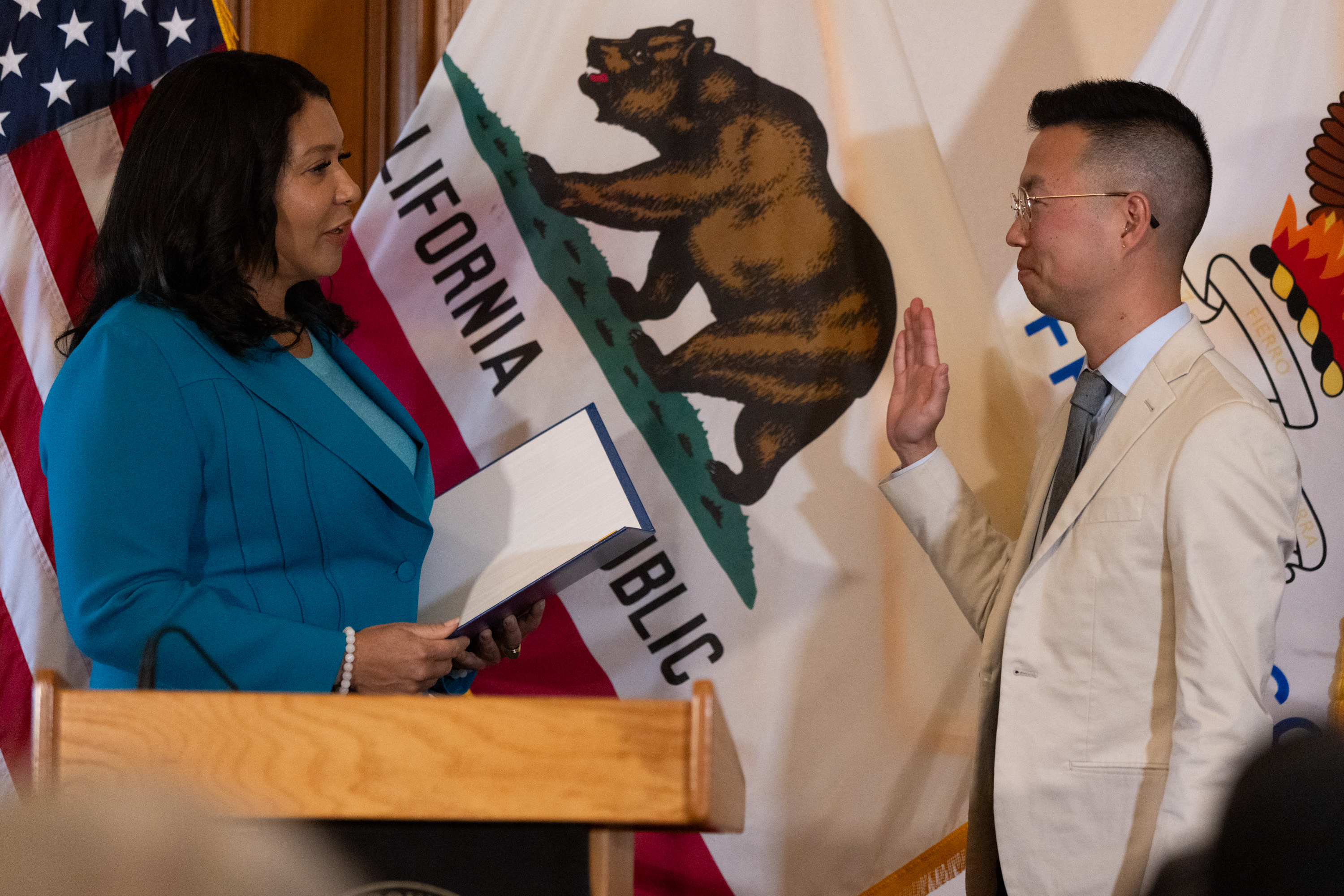 A woman in a teal suit holds a book, standing before a man in a beige suit who raises his right hand. Both stand in front of California and U.S. flags.