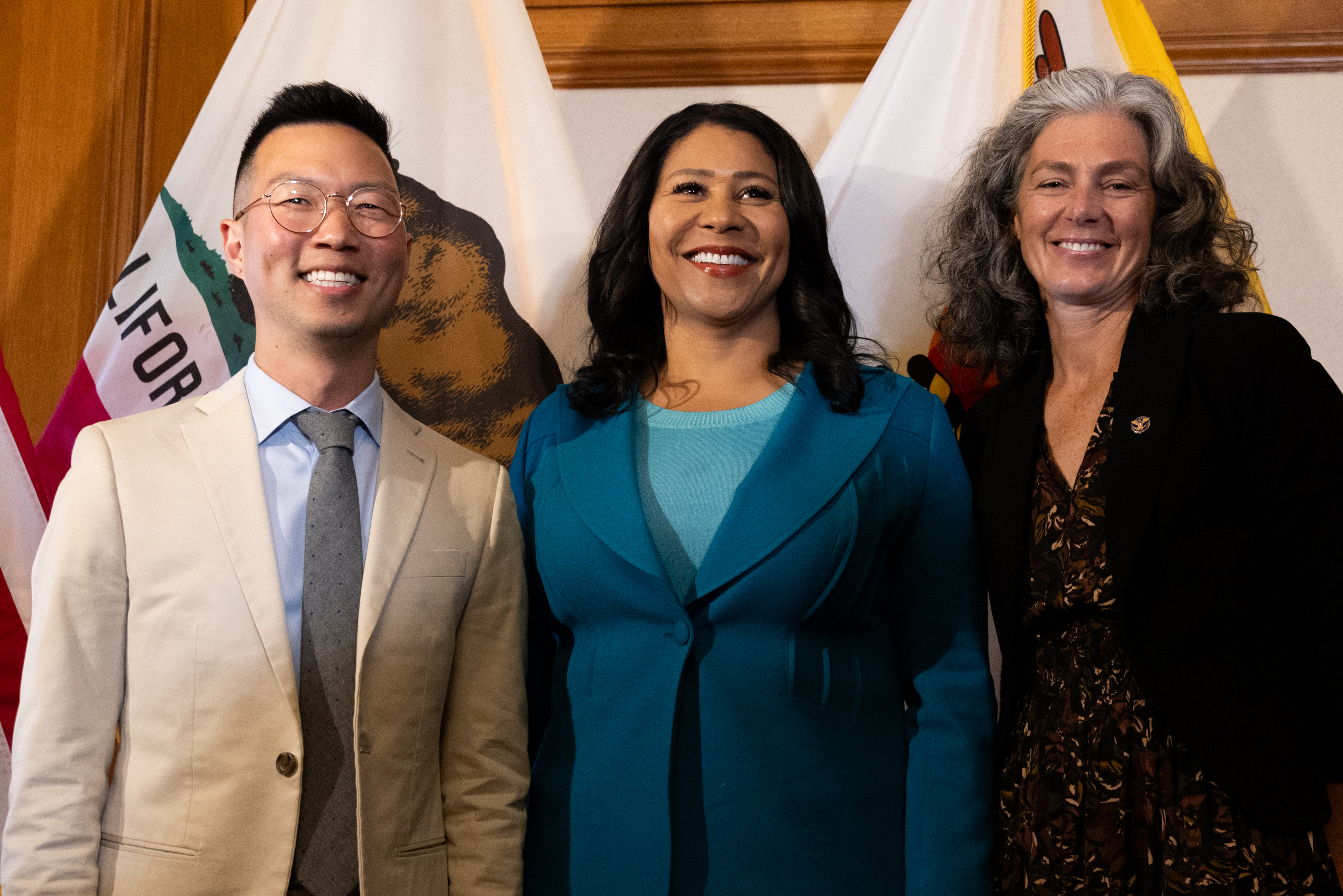 Three people are posing and smiling in front of California flags: a man in a beige suit, a woman in a turquoise jacket, and a woman in a black blazer.