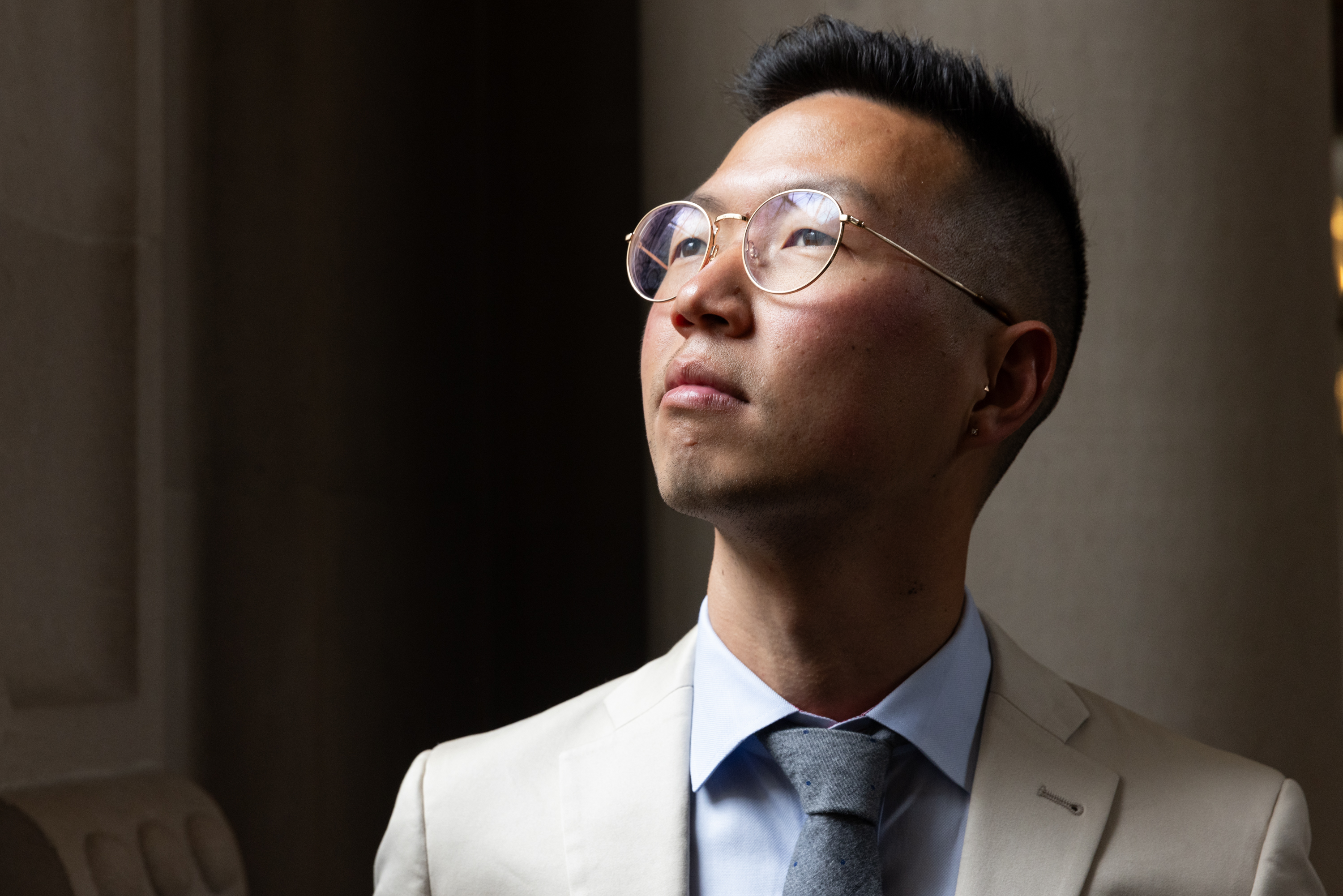 A person wearing glasses and a light-colored suit looks upward thoughtfully in soft lighting, with a dark background providing contrast.