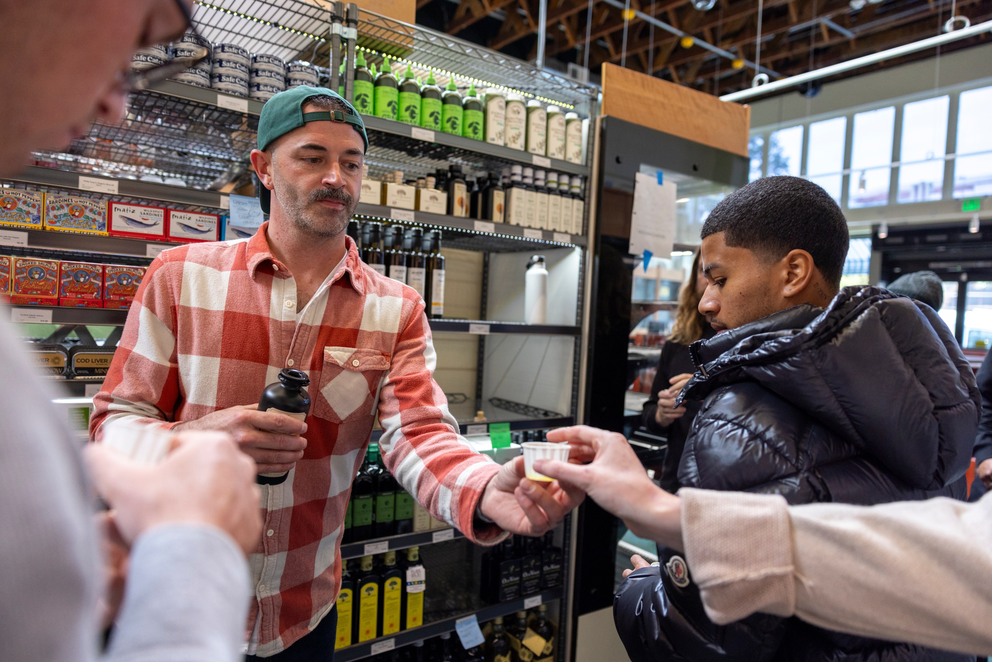 A man in a checkered shirt offers a small cup to another person in a store. Shelves behind them hold various bottles and canned goods.