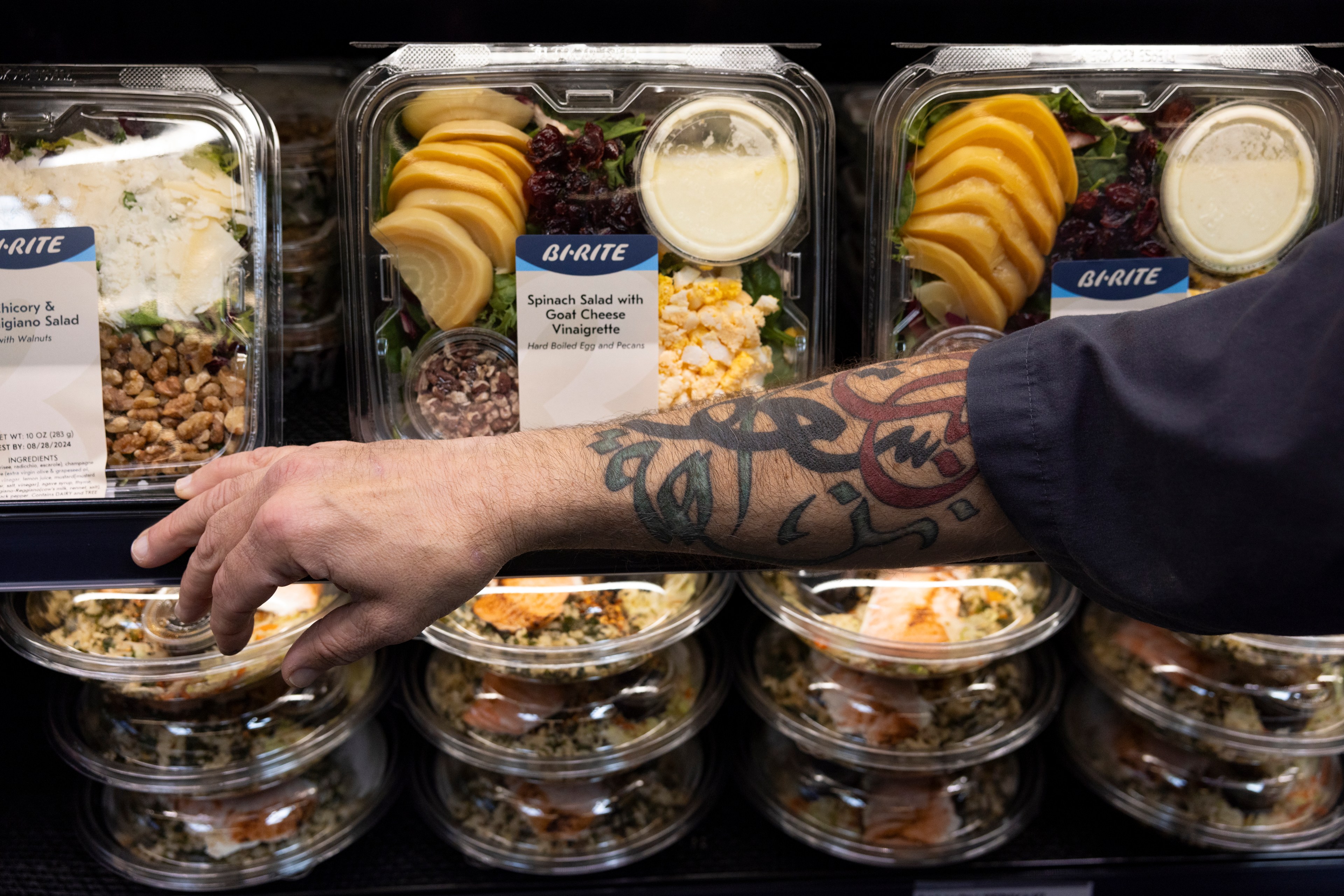 A tattooed arm reaches for a pre-packaged salad from a refrigerated shelf. The salads are labeled as &quot;Spinach Salad with Goat Cheese&quot; and &quot;Chicory &amp; Parmigiano Salad.&quot;