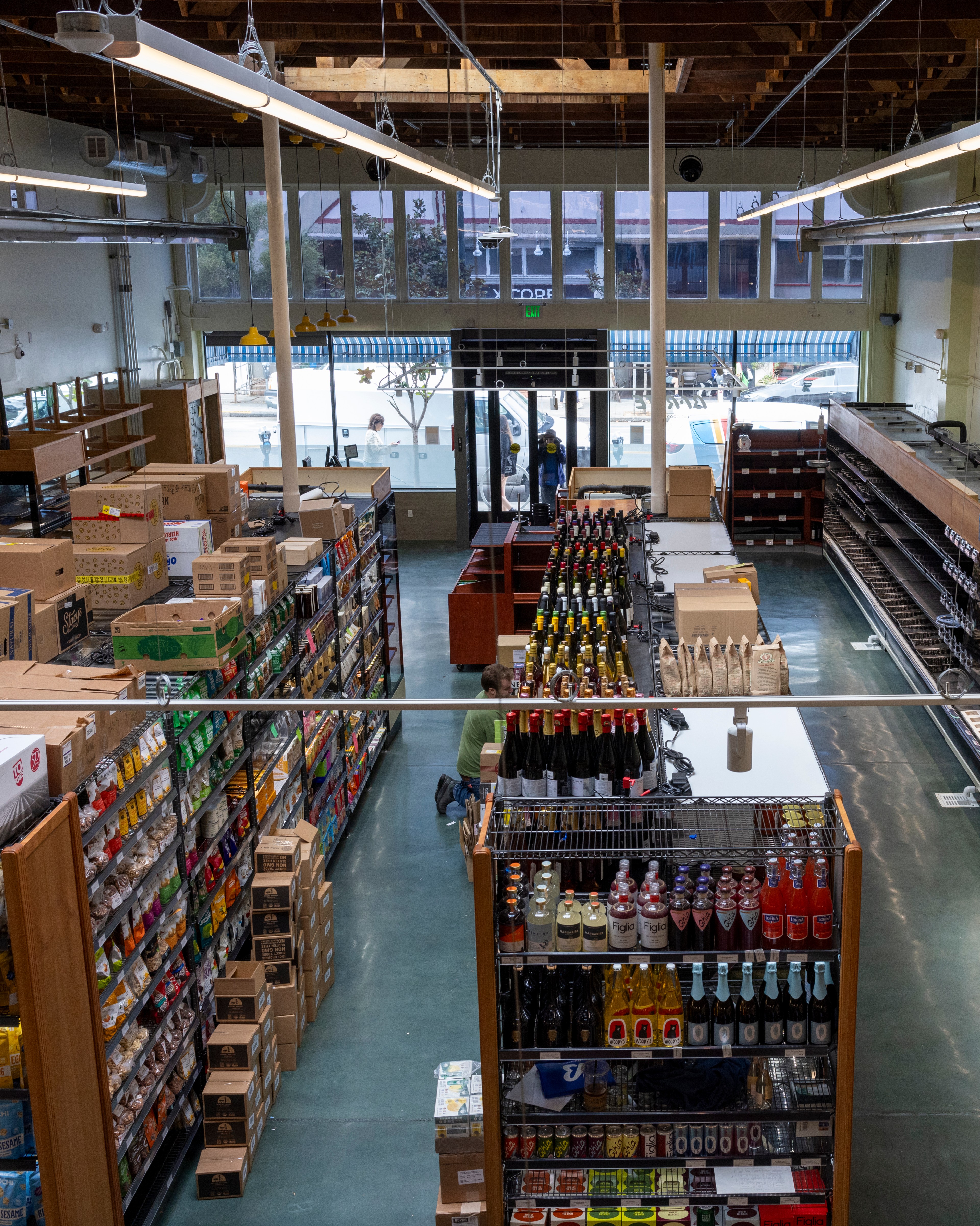 The image shows an organized grocery store's interior with shelves packed with various products, including beverages and snacks. A person kneels between aisles, likely restocking.