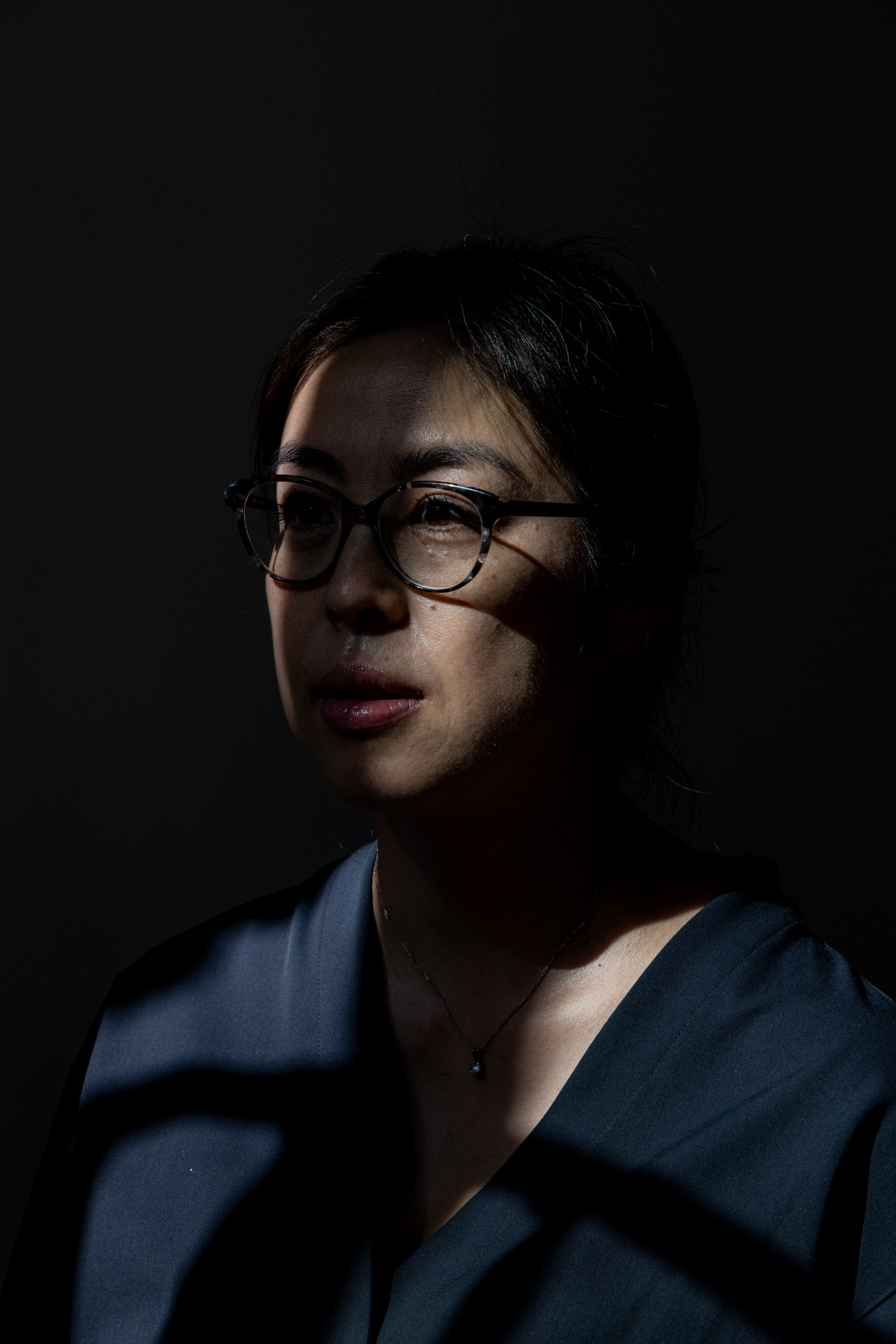 A woman wearing glasses and a dark shirt is partially illuminated by soft, directional lighting with shadows creating lines across her face.