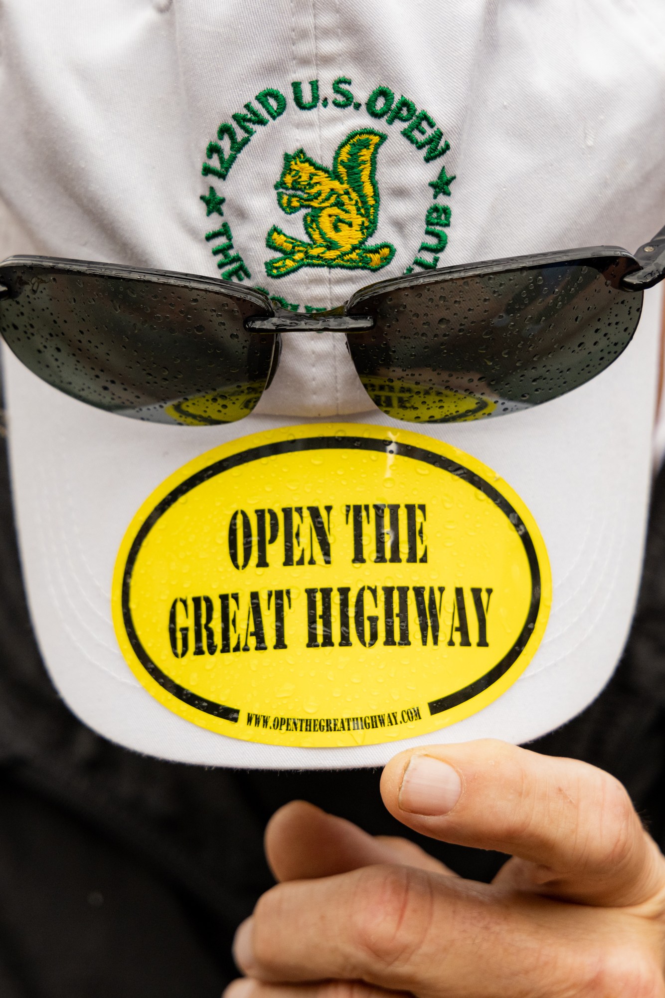 A person in a black jacket holds the brim of a white cap with dark sunglasses attached. The cap displays a yellow sticker reading &quot;OPEN THE GREAT HIGHWAY.&quot;