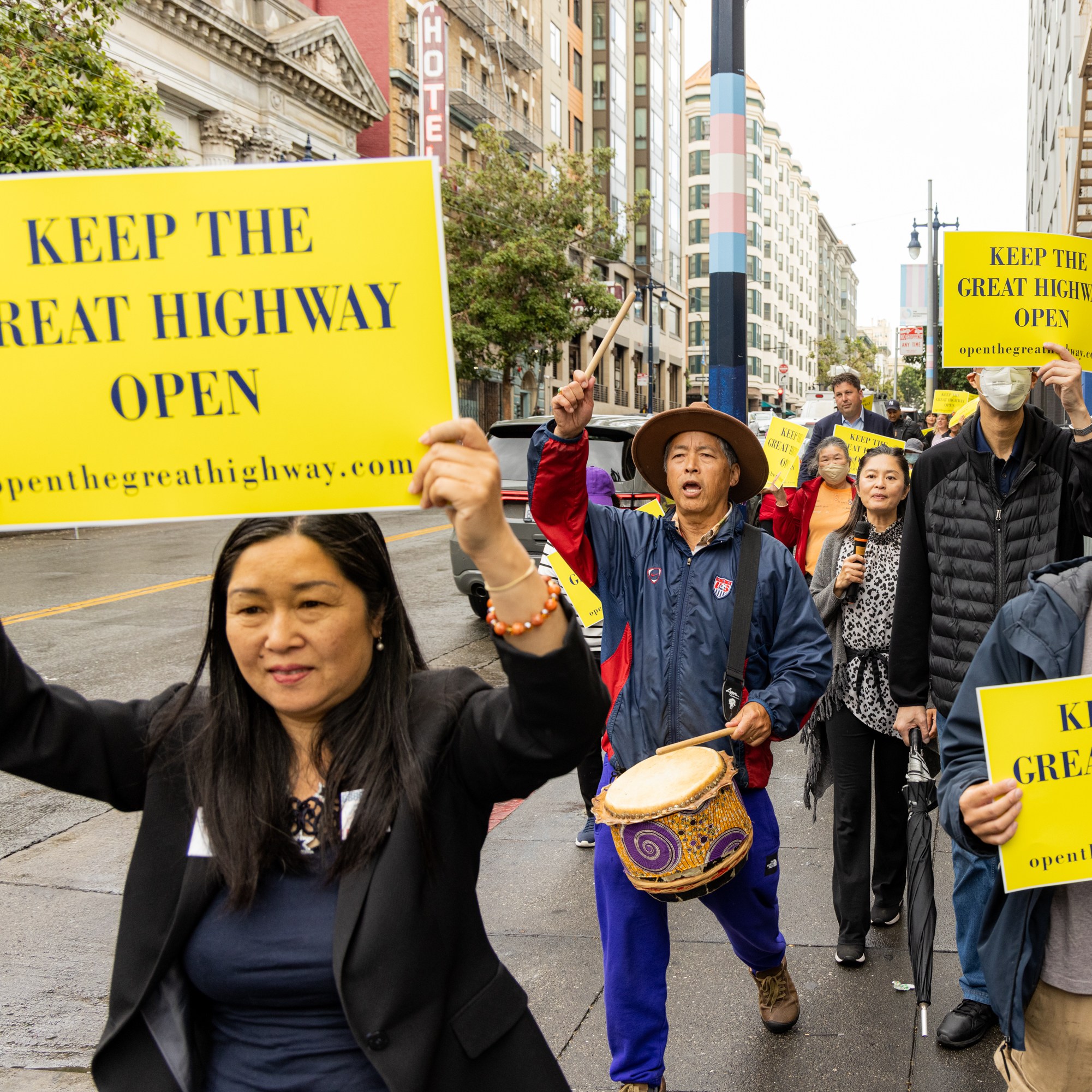 A group of people marches on a city street holding up yellow signs that read, &quot;KEEP THE GREAT HIGHWAY OPEN.&quot; One person is playing a drum.