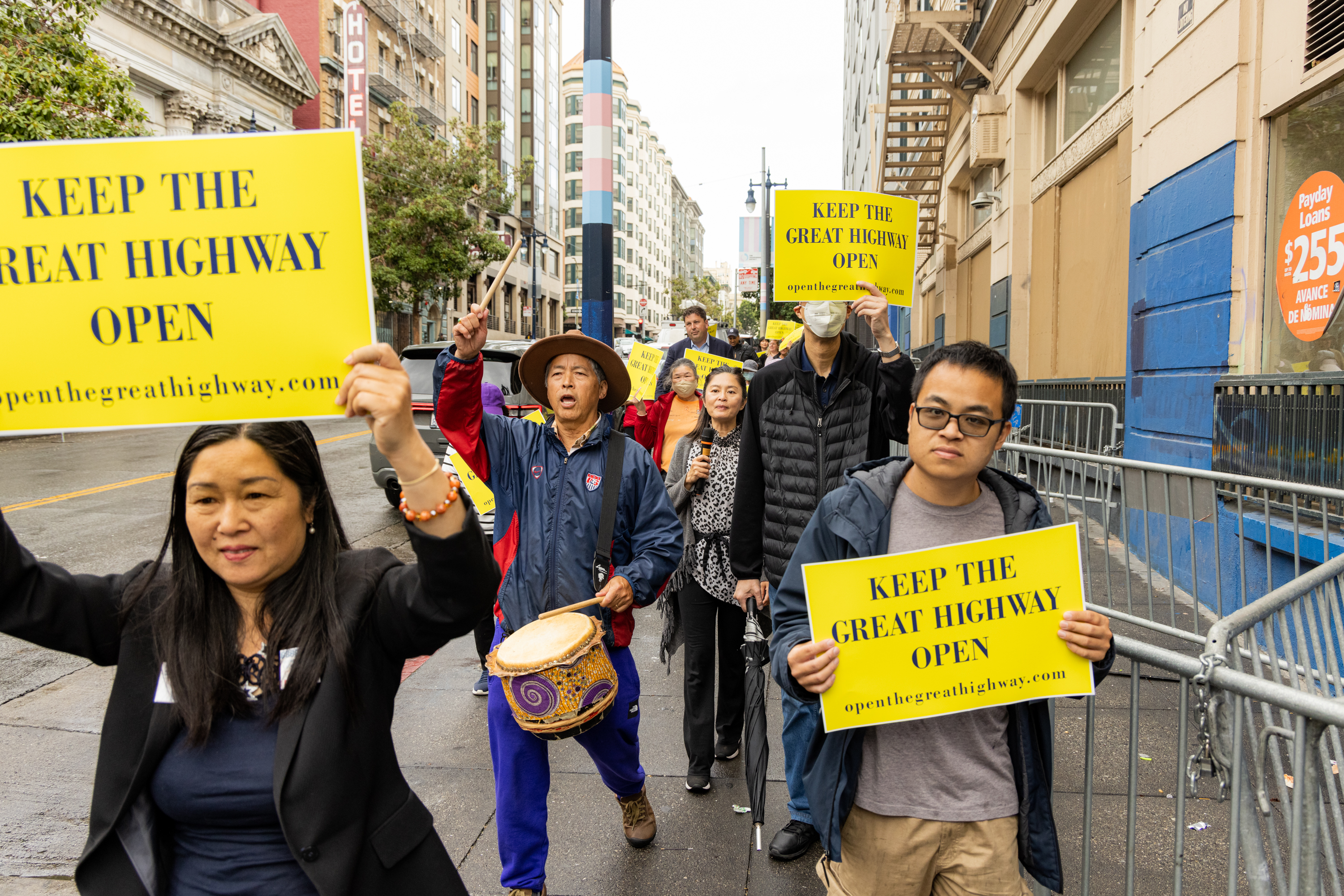 A group of people marches on a city street holding up yellow signs that read, &quot;KEEP THE GREAT HIGHWAY OPEN.&quot; One person is playing a drum.