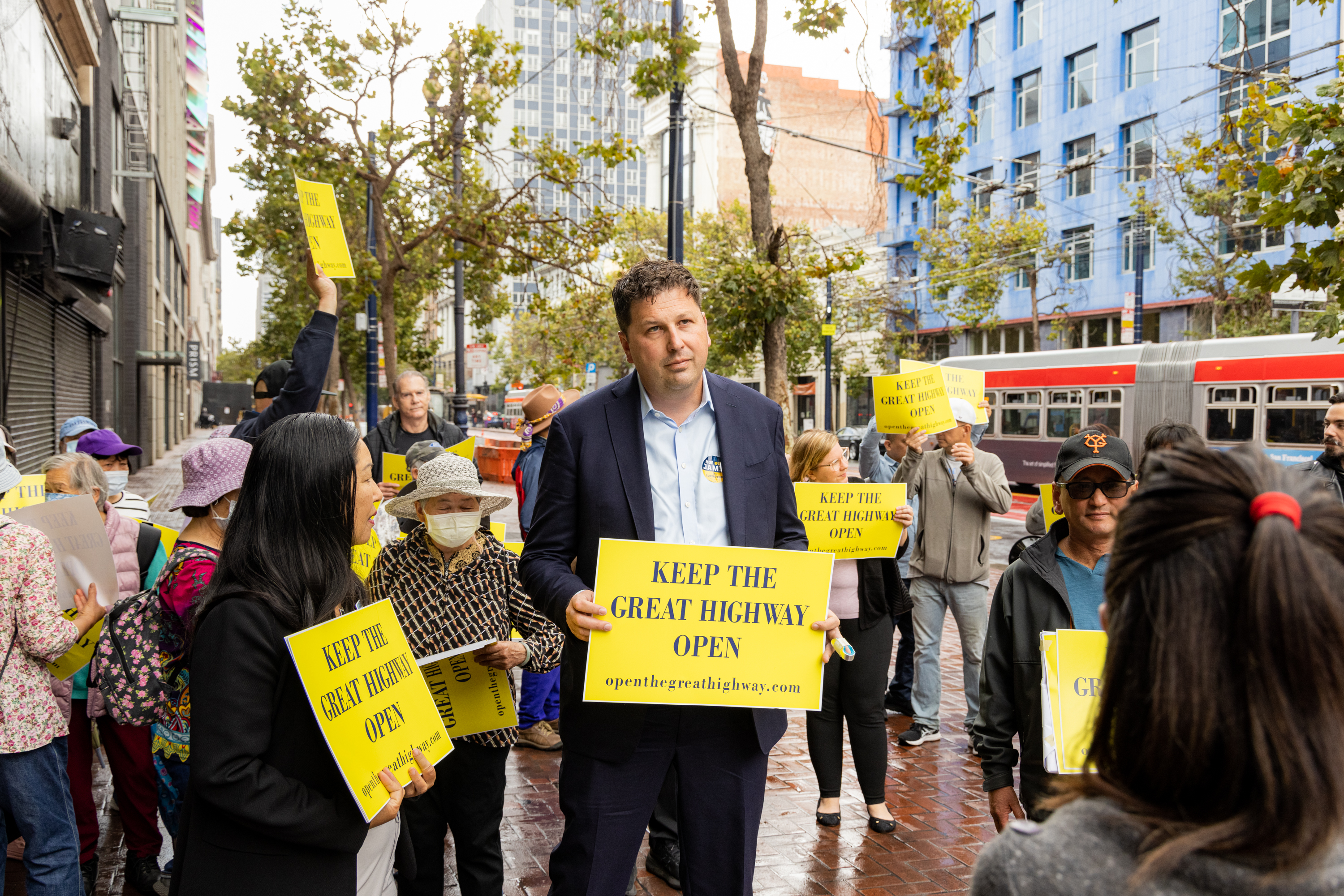 A group of people is standing on a city street holding yellow signs reading &quot;KEEP THE GREAT HIGHWAY OPEN.&quot; They appear to be participating in a protest or rally.