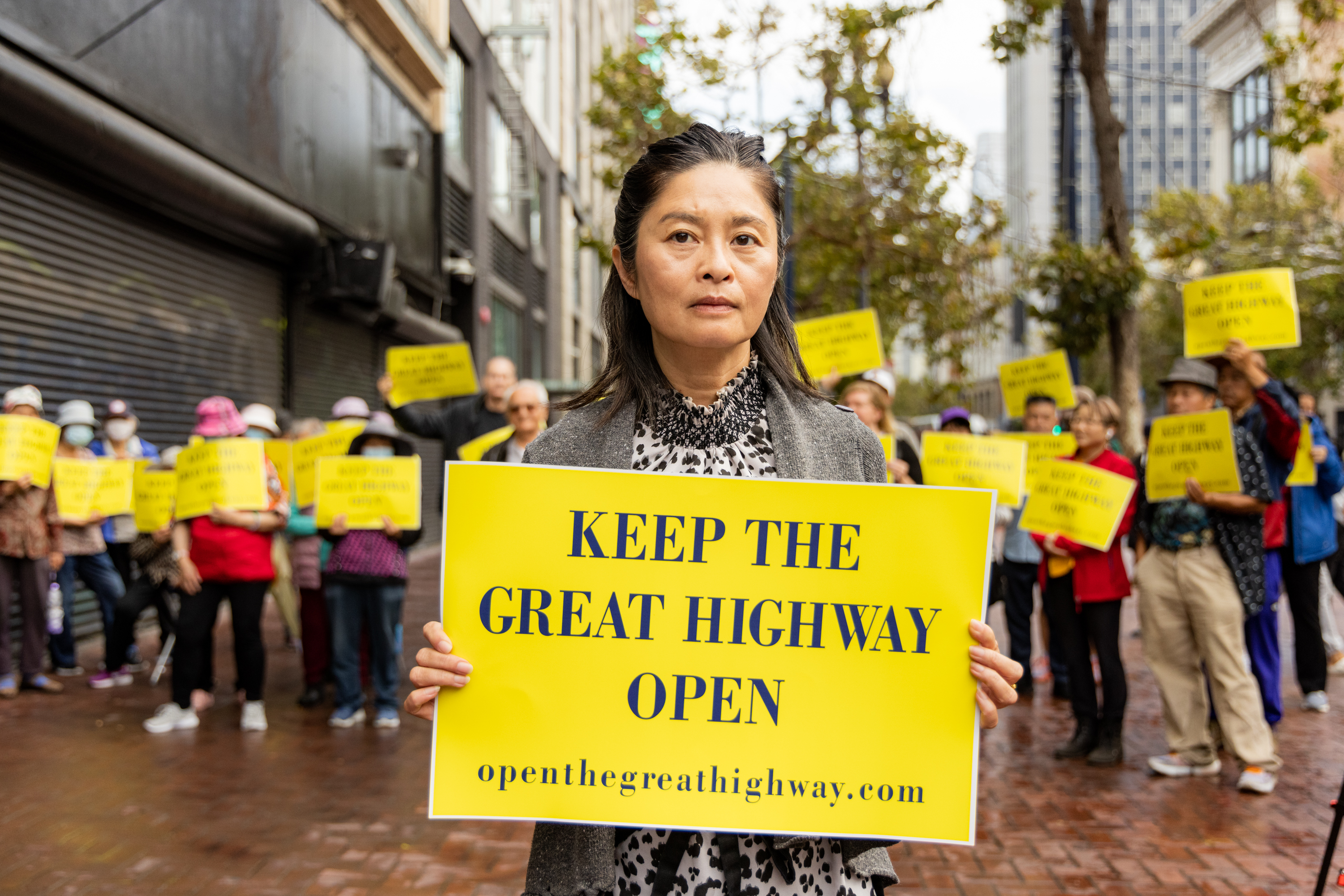 A woman stands in front of a crowd holding a bright yellow sign reading &quot;KEEP THE GREAT HIGHWAY OPEN.&quot; The crowd behind her holds similar signs.