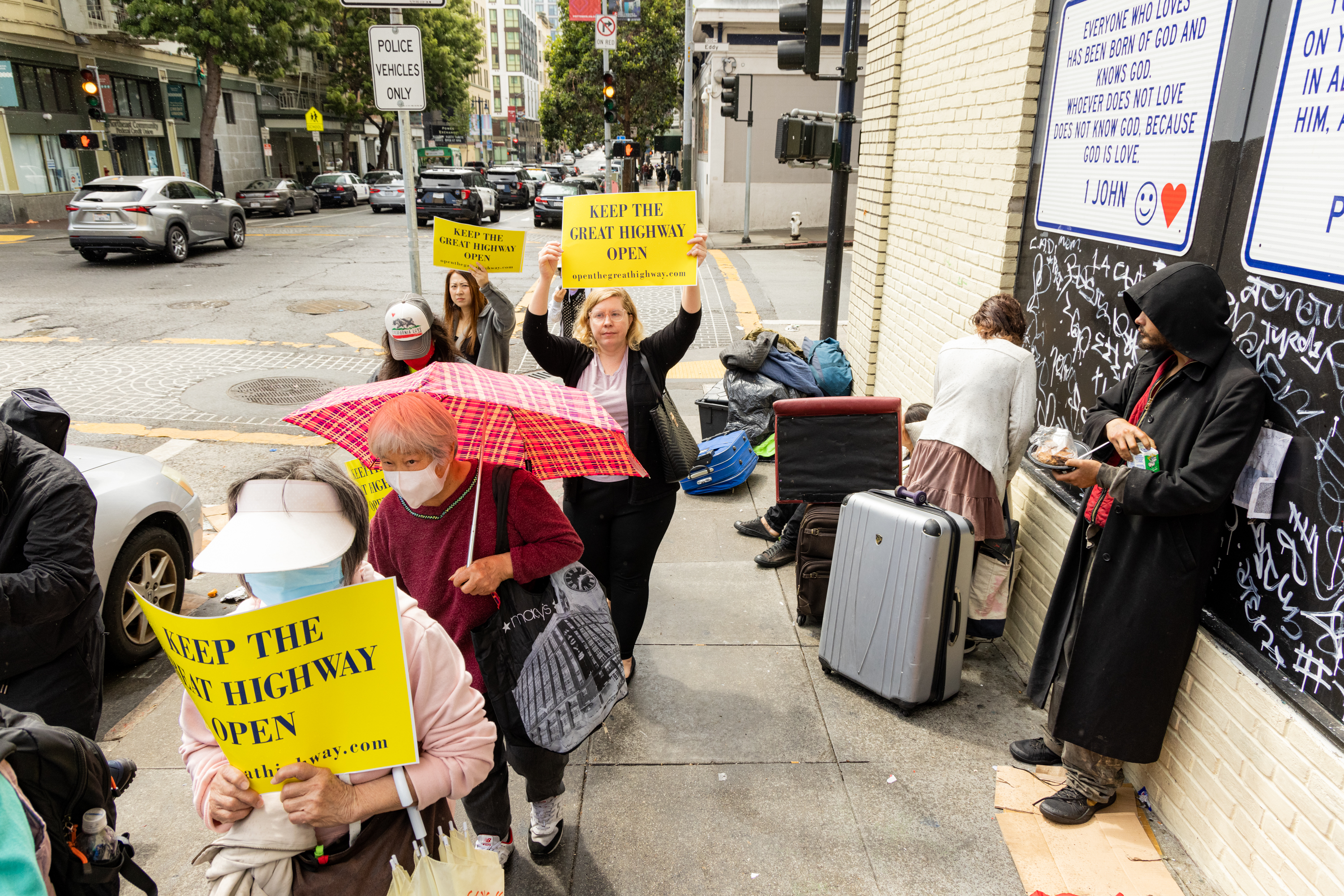 A group of people holding yellow signs reading &quot;KEEP THE GREAT HIGHWAY OPEN&quot; stand on a busy sidewalk. Nearby, there are individuals with suitcases resting or standing.