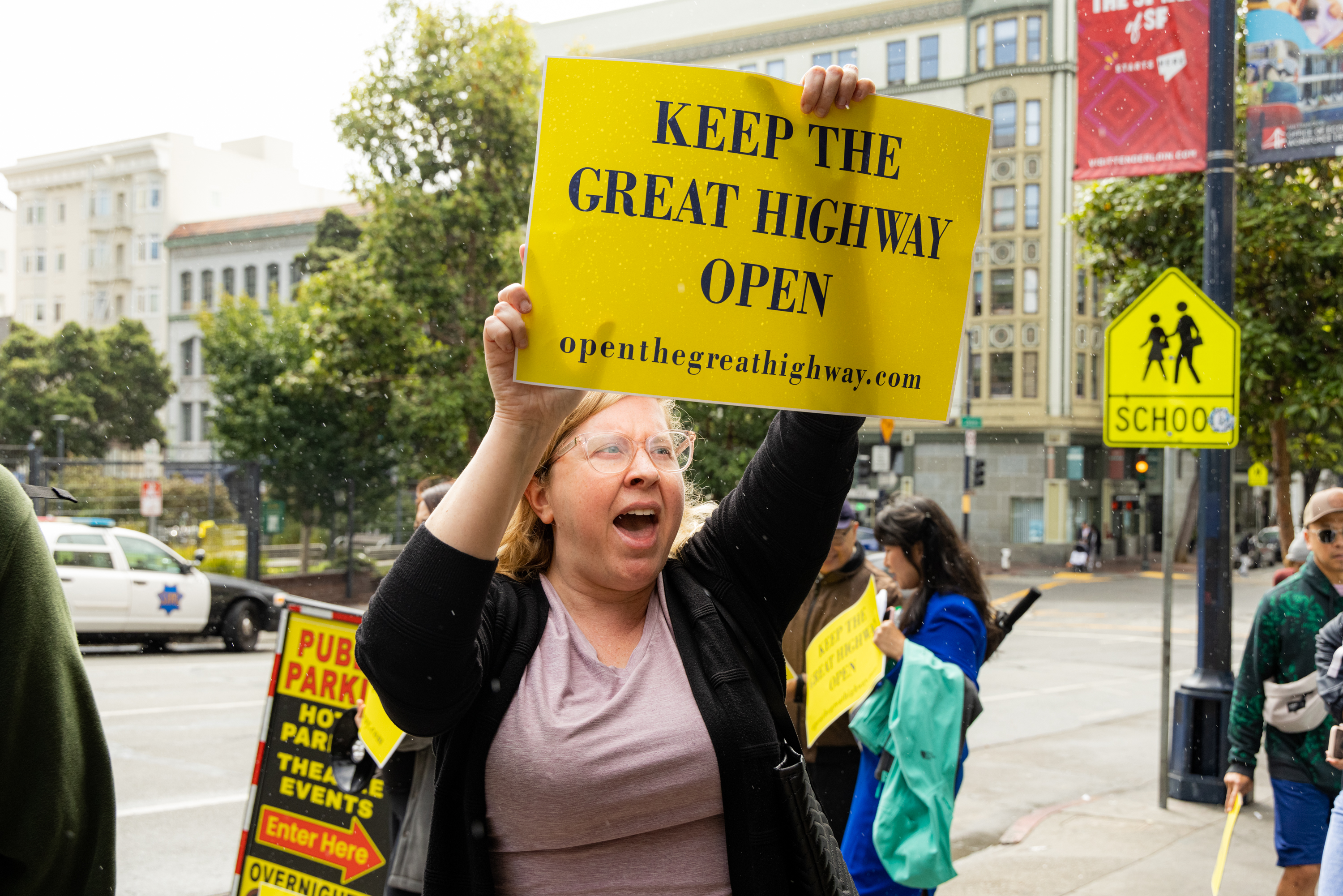 A woman enthusiastically holds a yellow sign reading, &quot;Keep the Great Highway Open&quot; in a city street with trees, buildings, and a school crossing sign visible in the background.