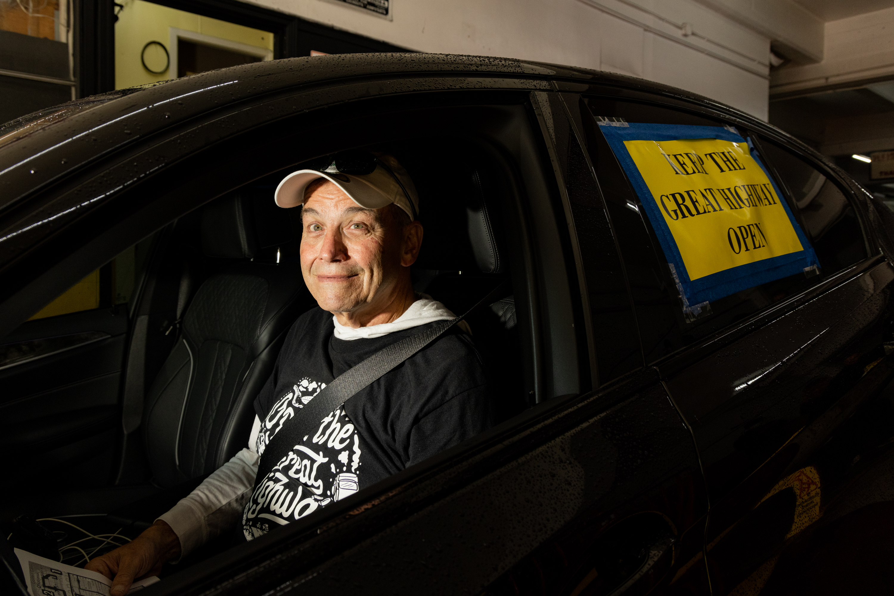 A man wearing a white baseball cap and a black shirt sits in the driver's seat of a car. The car window has a yellow sign reading &quot;KEEP THE GREAT HIGHWAY OPEN.&quot;