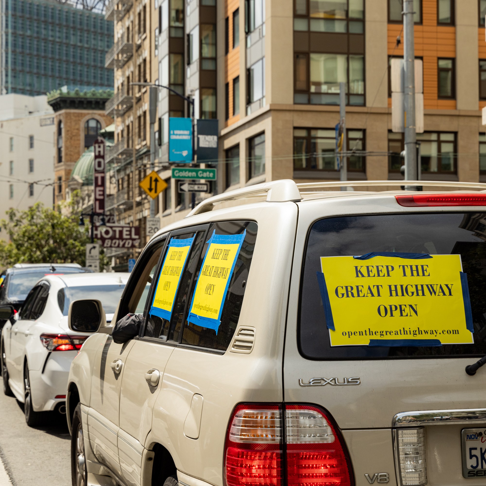 In a city with tall buildings, cars are lined up on the street. The leading car, a Lexus, has yellow signs saying &quot;KEEP THE GREAT HIGHWAY OPEN&quot; taped to its windows.
