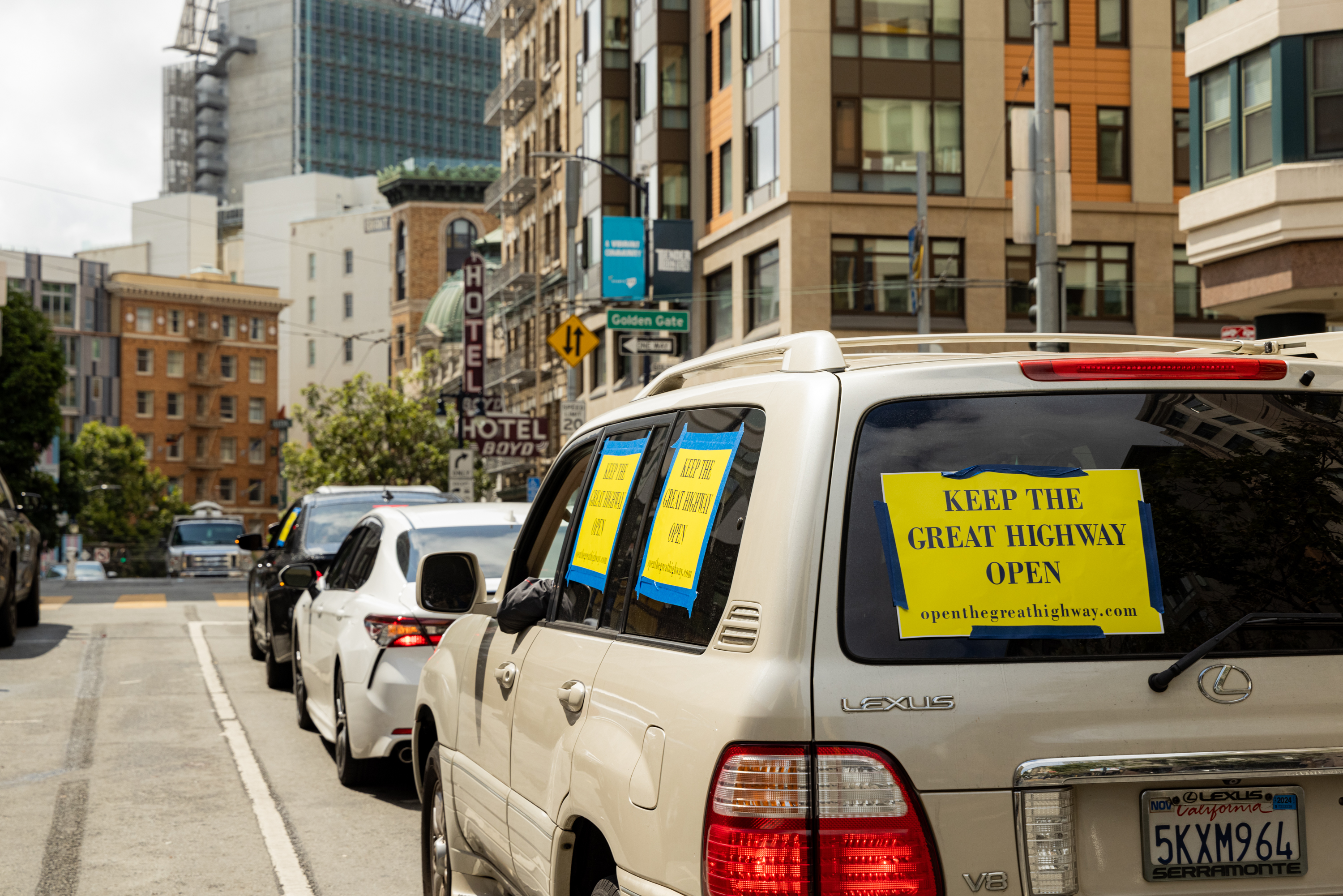 In a city with tall buildings, cars are lined up on the street. The leading car, a Lexus, has yellow signs saying &quot;KEEP THE GREAT HIGHWAY OPEN&quot; taped to its windows.