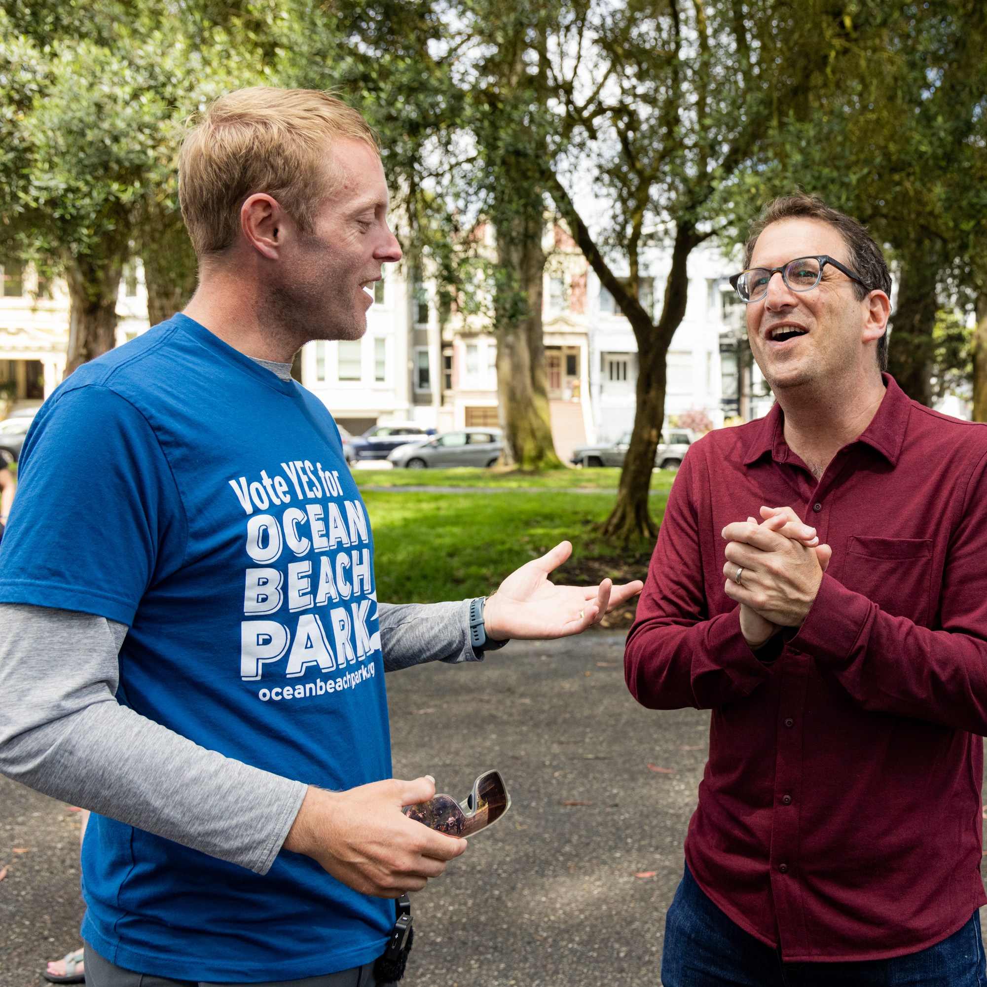 Two men conversing in a park, one in a blue &quot;Vote YES for Ocean Beach Park&quot; shirt, the other in a red shirt, while another man in a similar blue shirt walks behind them.