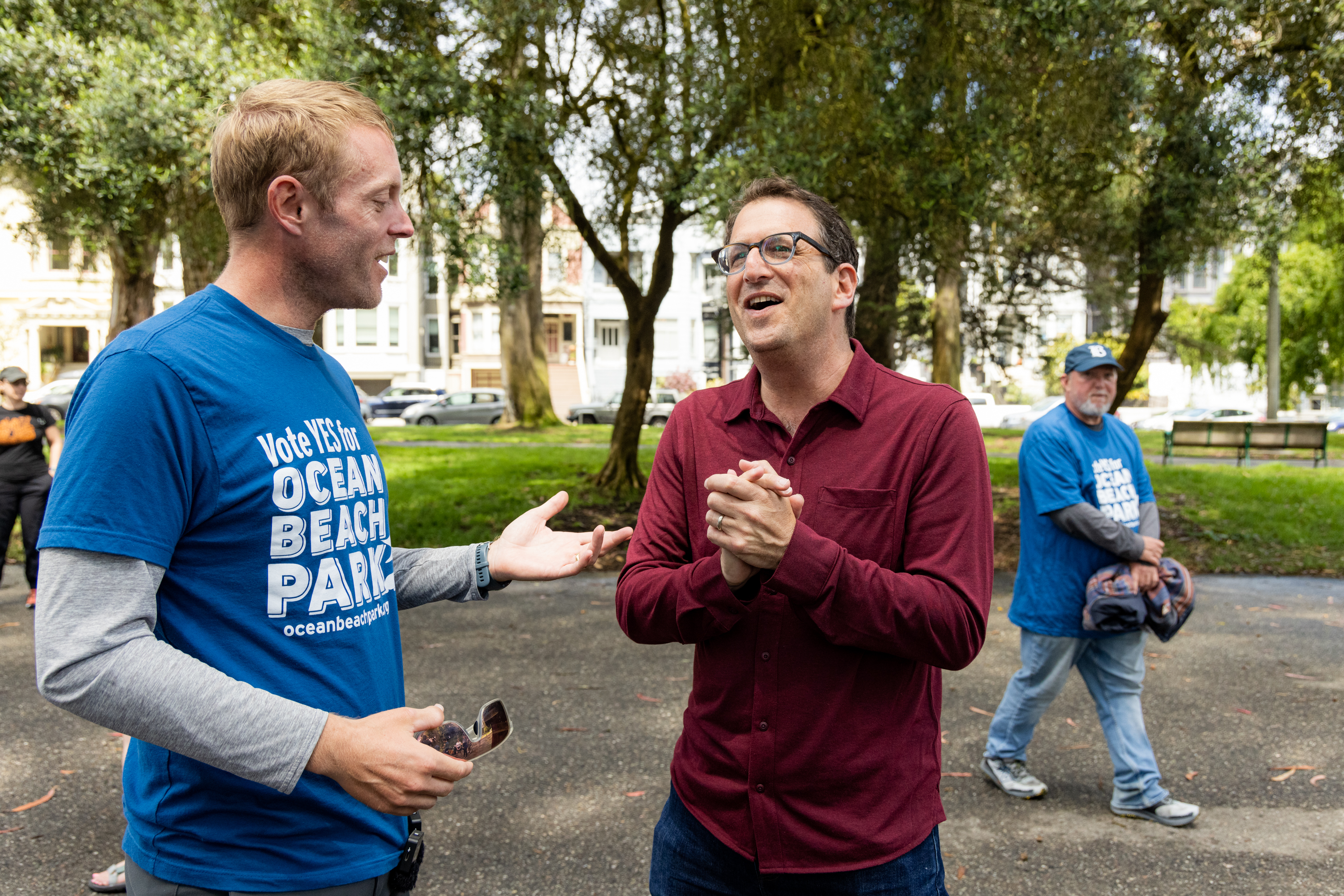 Two men conversing in a park, one in a blue &quot;Vote YES for Ocean Beach Park&quot; shirt, the other in a red shirt, while another man in a similar blue shirt walks behind them.