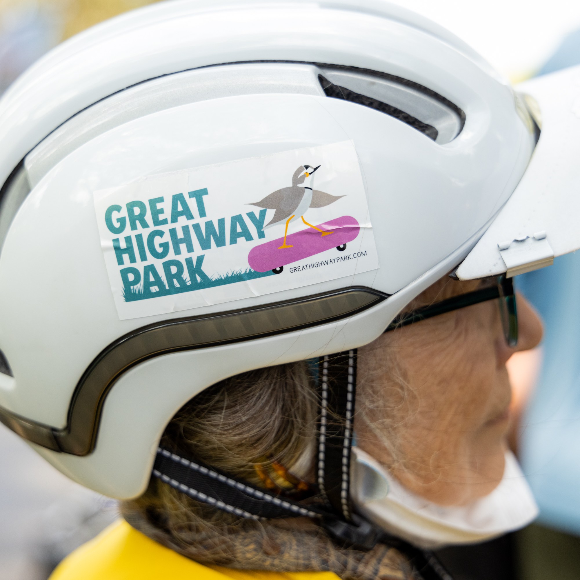 An individual wearing a white helmet with a sticker that reads &quot;Great Highway Park&quot; and depicts a bird on a skateboard.