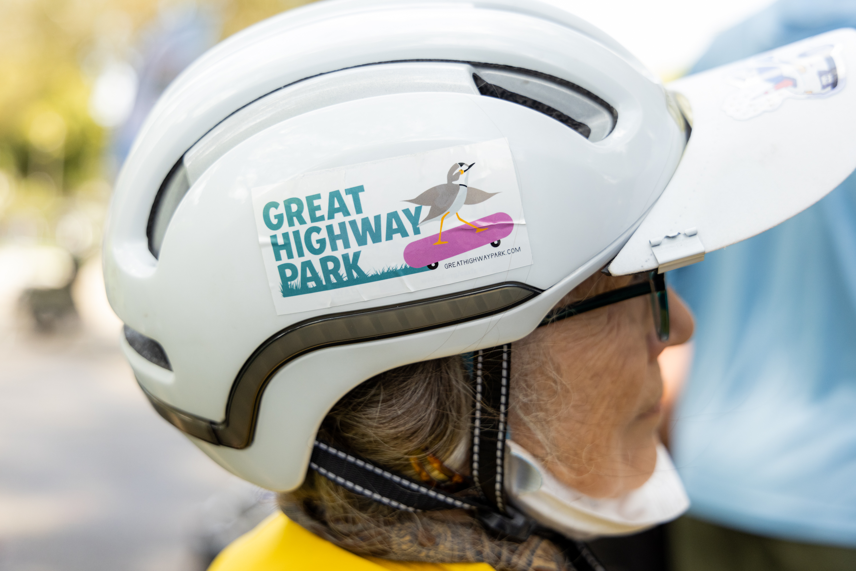 An individual wearing a white helmet with a sticker that reads &quot;Great Highway Park&quot; and depicts a bird on a skateboard.