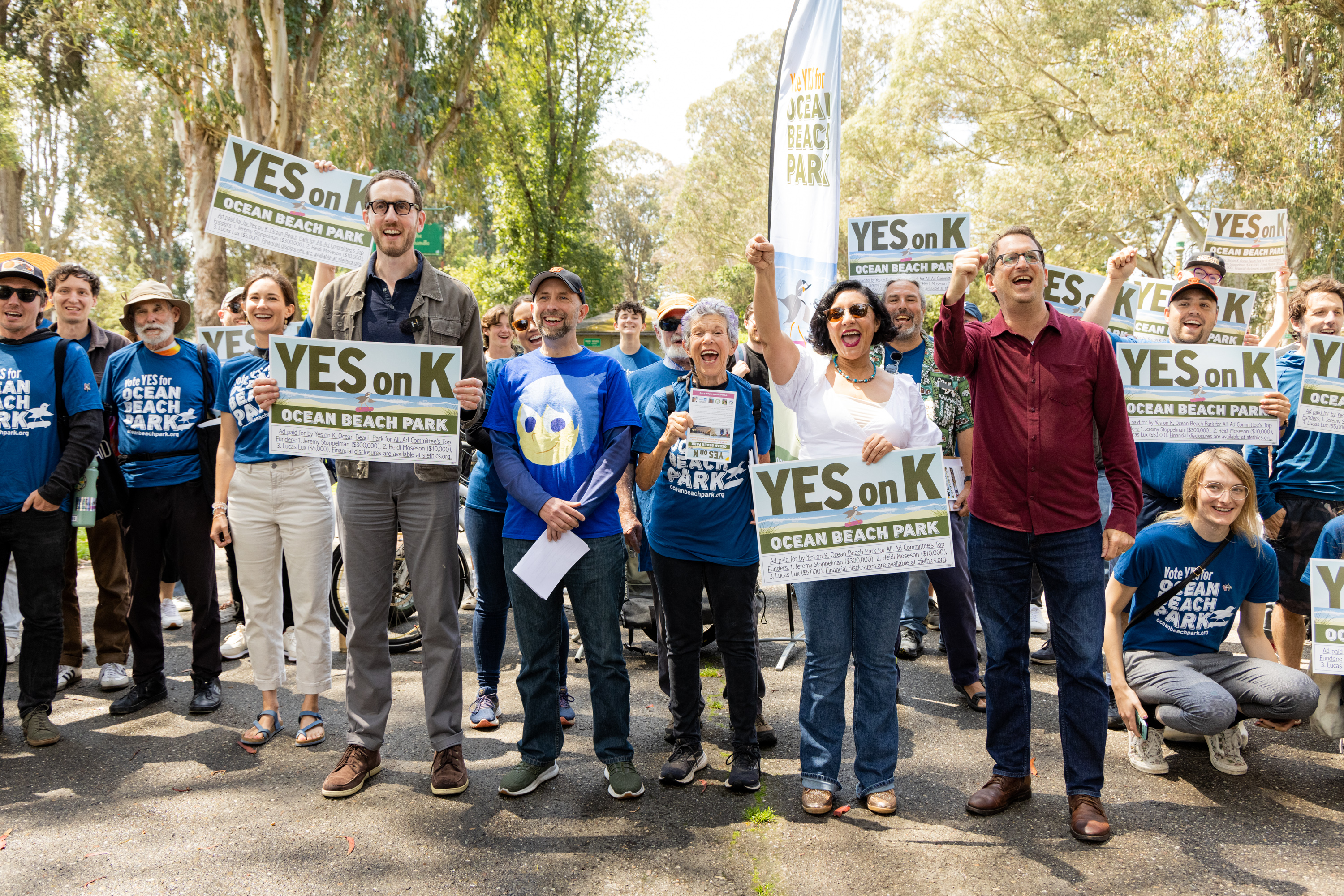 A group of enthusiastic people outdoors hold signs that say &quot;YES on K&quot; advocating for Ocean Beach Park, smiling and raising their hands in support.