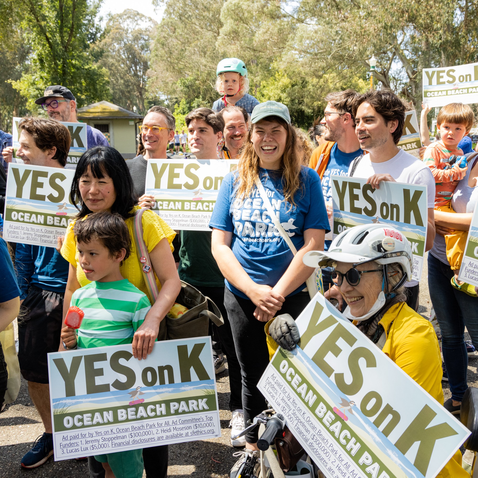 A diverse group of people, including children, stand outdoors holding signs that say &quot;YES on K&quot; and &quot;OCEAN BEACH PARK.&quot; They appear to be participating in a rally or gathering.