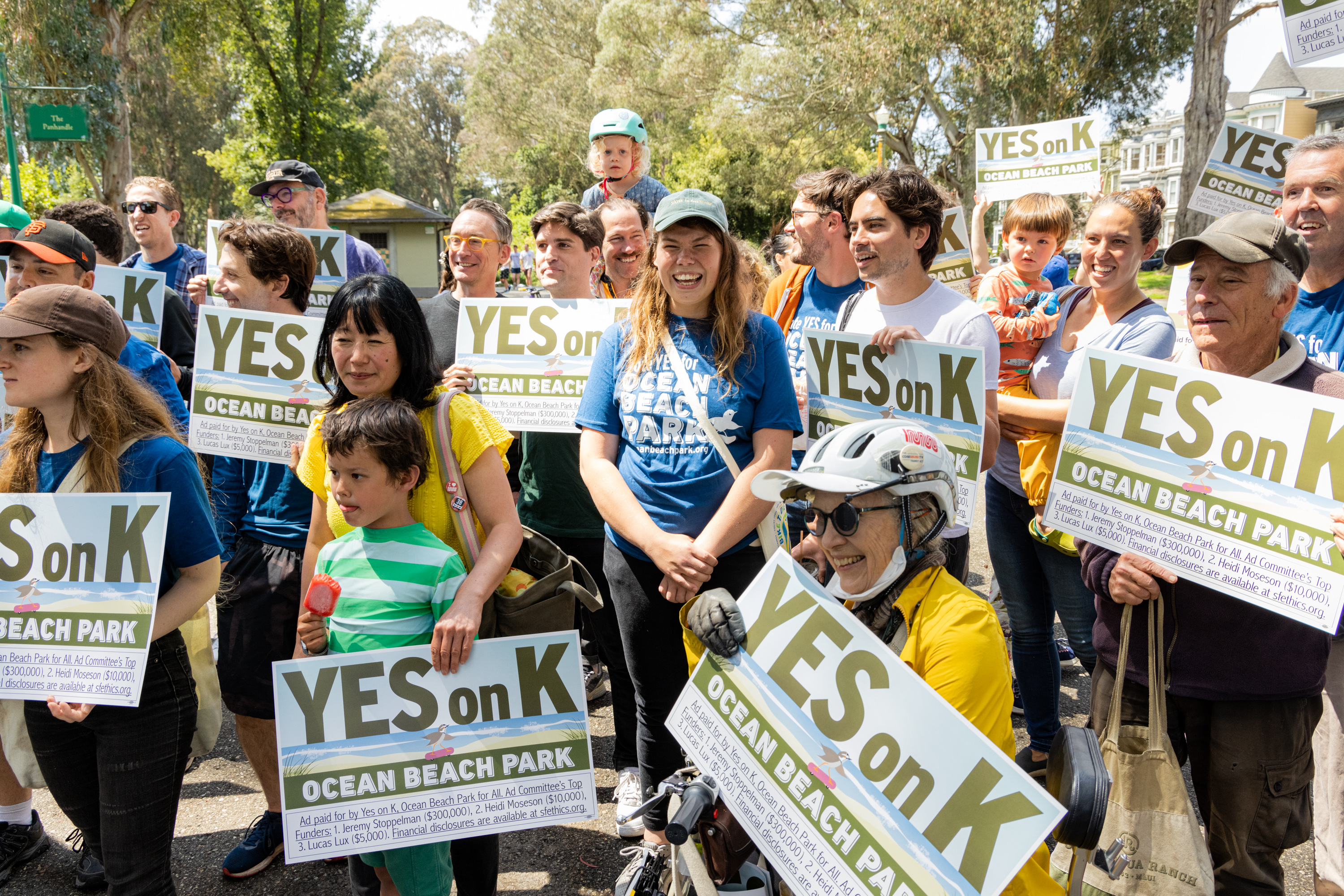 A diverse group of people, including children, stand outdoors holding signs that say &quot;YES on K&quot; and &quot;OCEAN BEACH PARK.&quot; They appear to be participating in a rally or gathering.