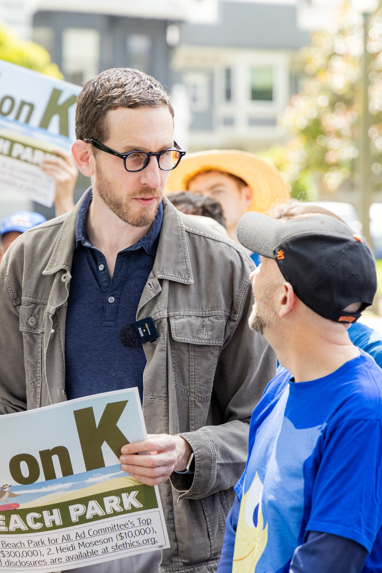 A group of people, some holding “YES on K” signs for Ocean Beach Park, are gathered outdoors. One person in a jacket and glasses is speaking to another wearing a cap.
