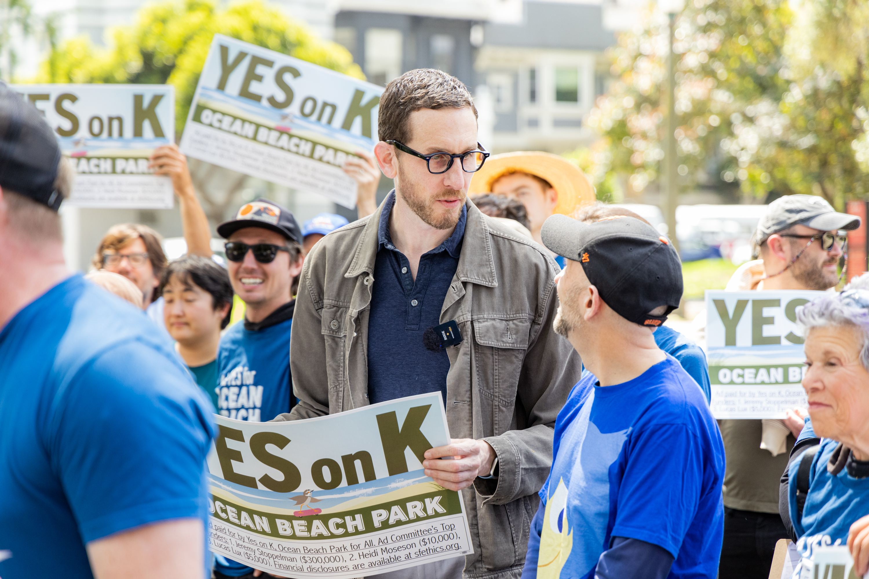 A group of people, some holding “YES on K” signs for Ocean Beach Park, are gathered outdoors. One person in a jacket and glasses is speaking to another wearing a cap.