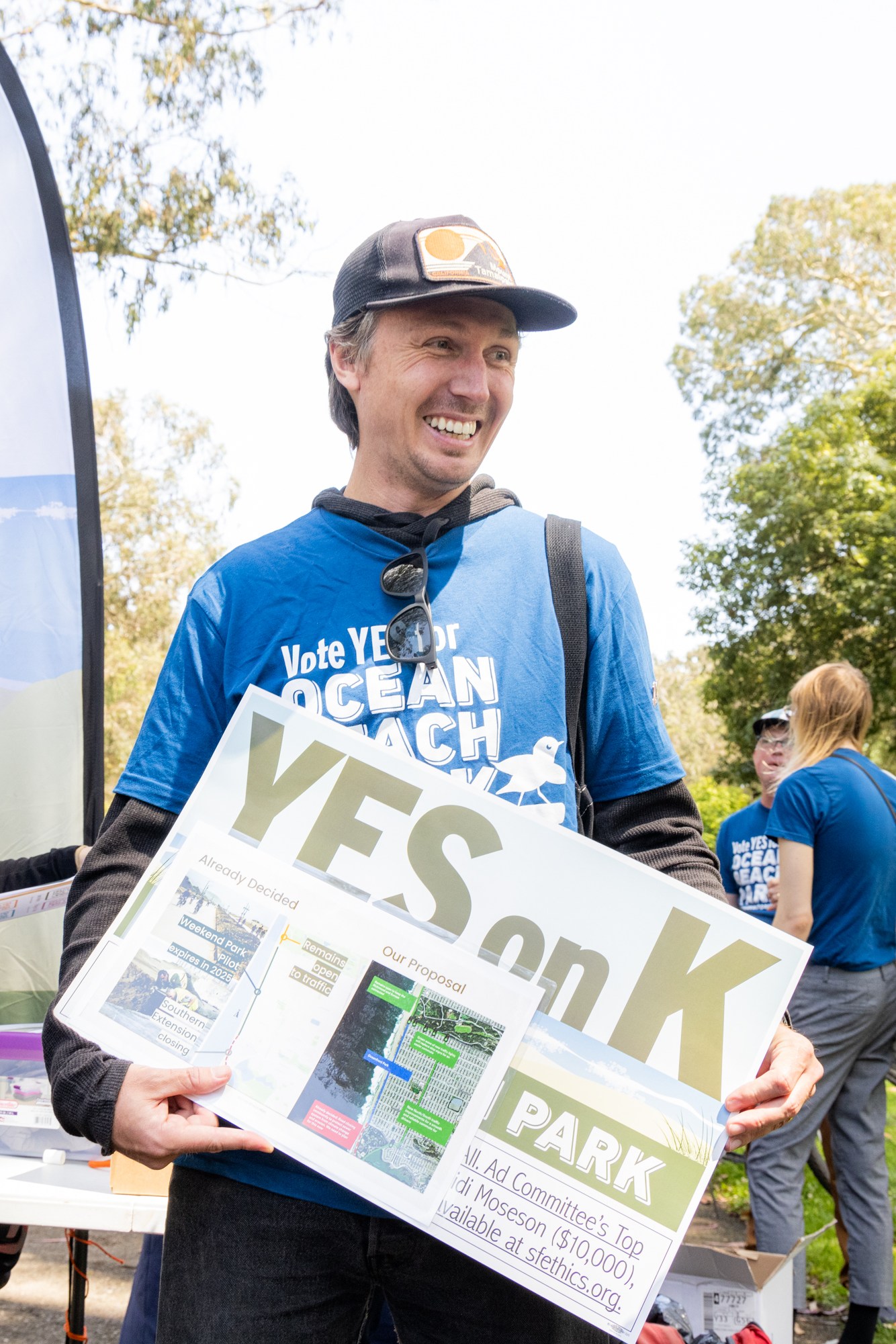 People wearing blue "Vote YES for Ocean Beach Park" shirts are gathered, holding signs supporting Measure K at an outdoor event, with trees and banners in the background.