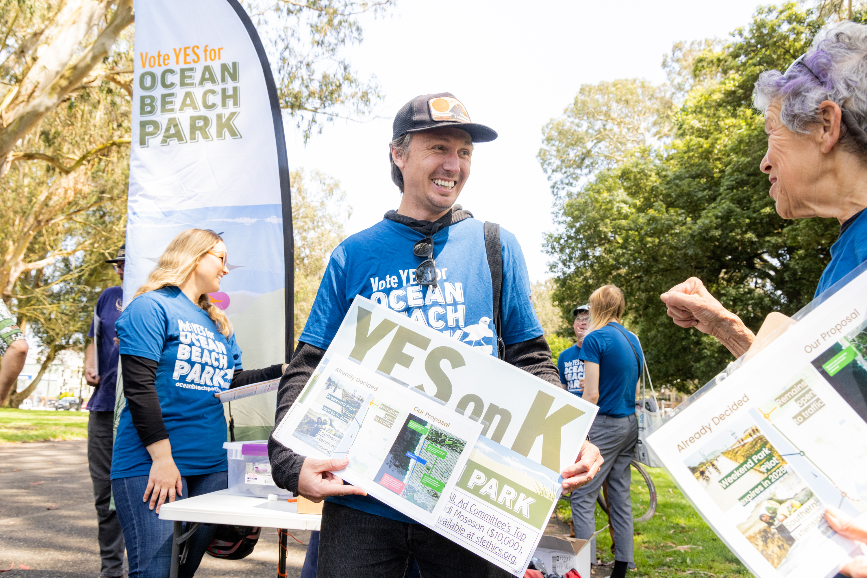 People wearing blue "Vote YES for Ocean Beach Park" shirts are gathered, holding signs supporting Measure K at an outdoor event, with trees and banners in the background.