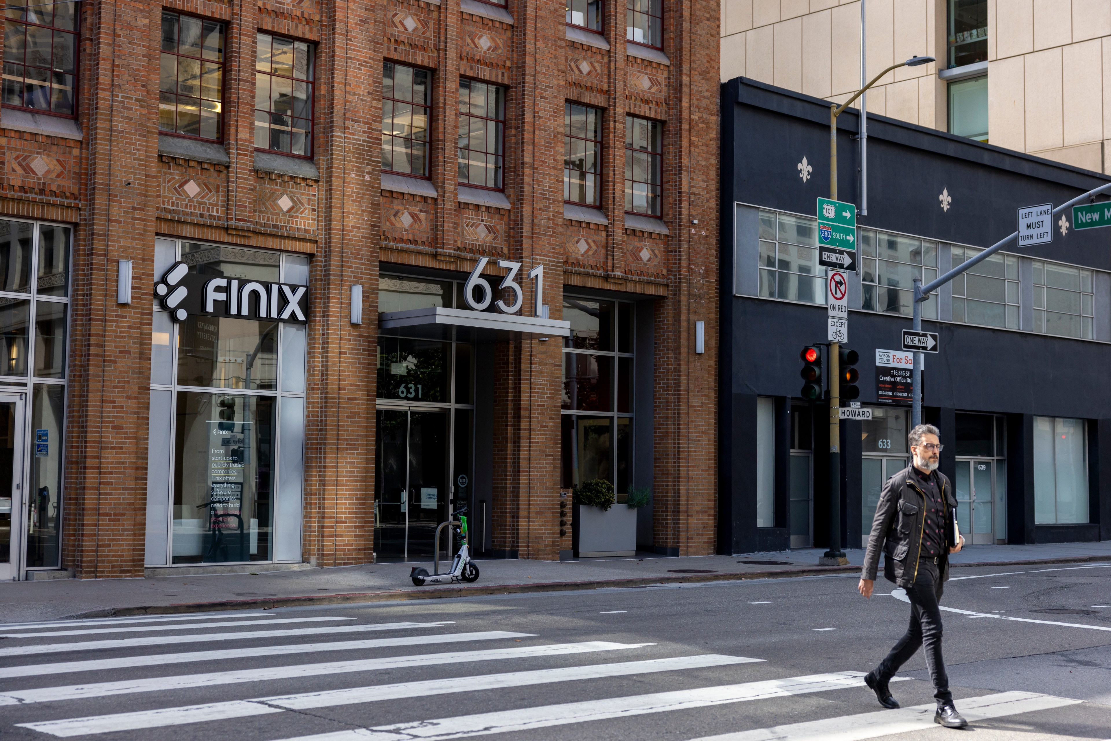 A man in a black jacket crosses a city street at a crosswalk. Behind him is a brick building with &quot;FINIX&quot; signage and the number &quot;631&quot; displayed above the entrance.