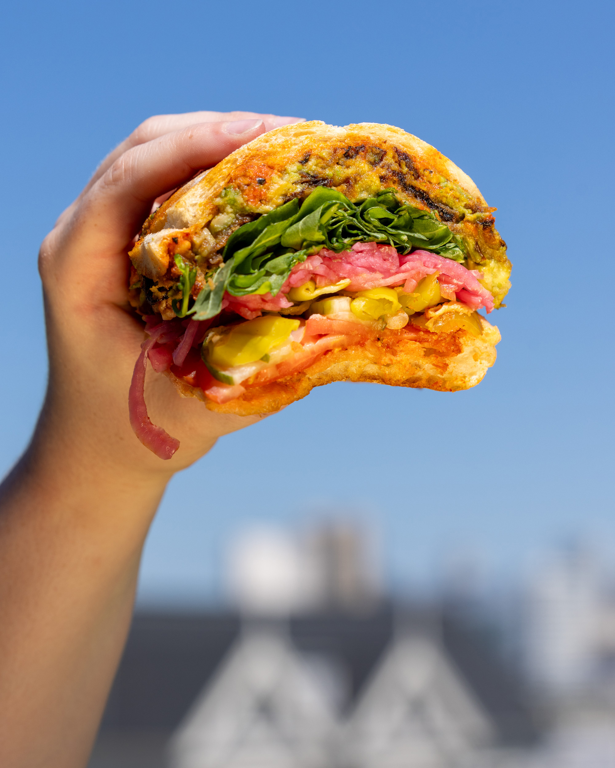 A hand holds a partially eaten sandwich with layers of meats, greens, and vegetables against a clear blue sky background.