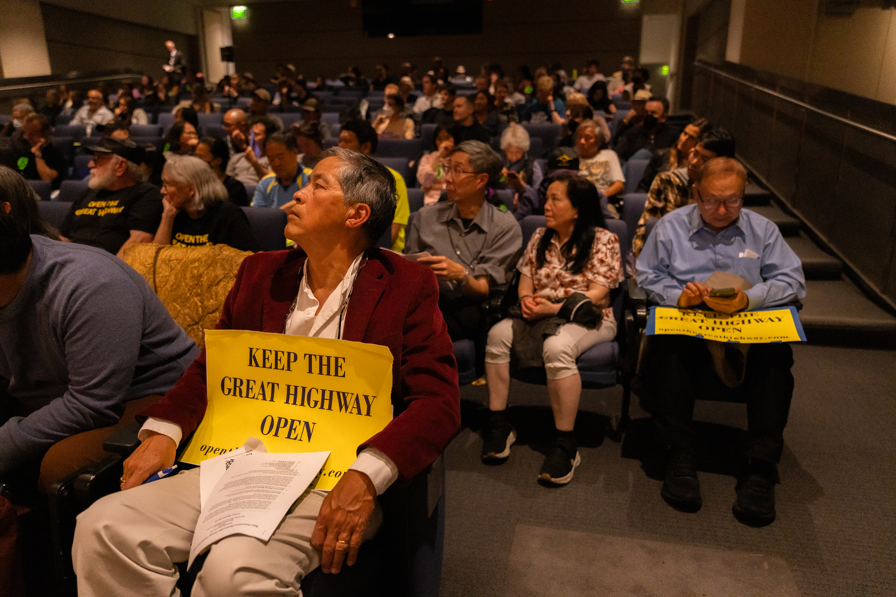 A seated crowd in a dimly lit auditorium listens attentively, with some holding yellow signs reading, &quot;KEEP THE GREAT HIGHWAY OPEN.&quot;