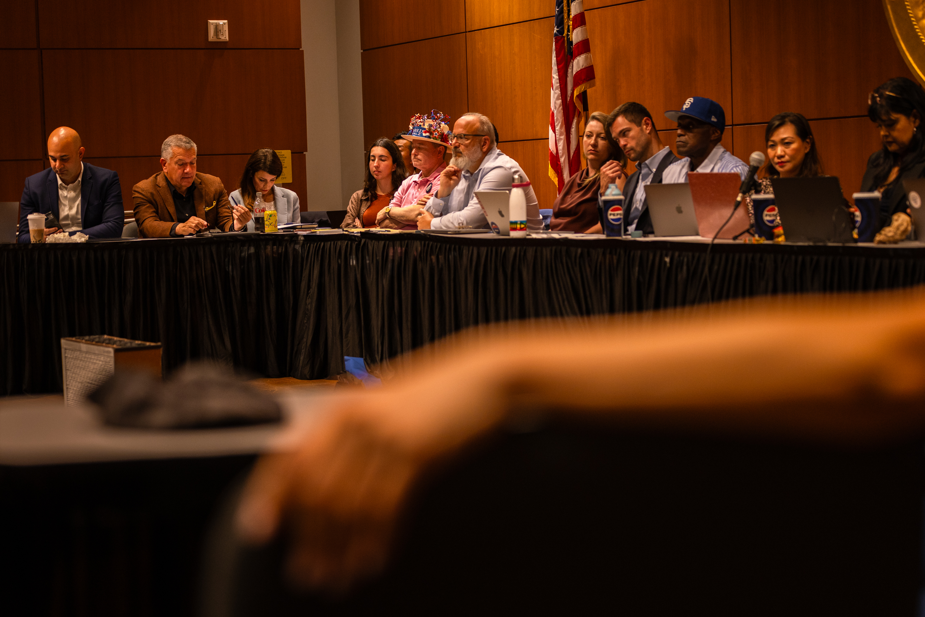 A group of people is seated at a long table in a conference room, each with laptops and papers, with an American flag in the background.