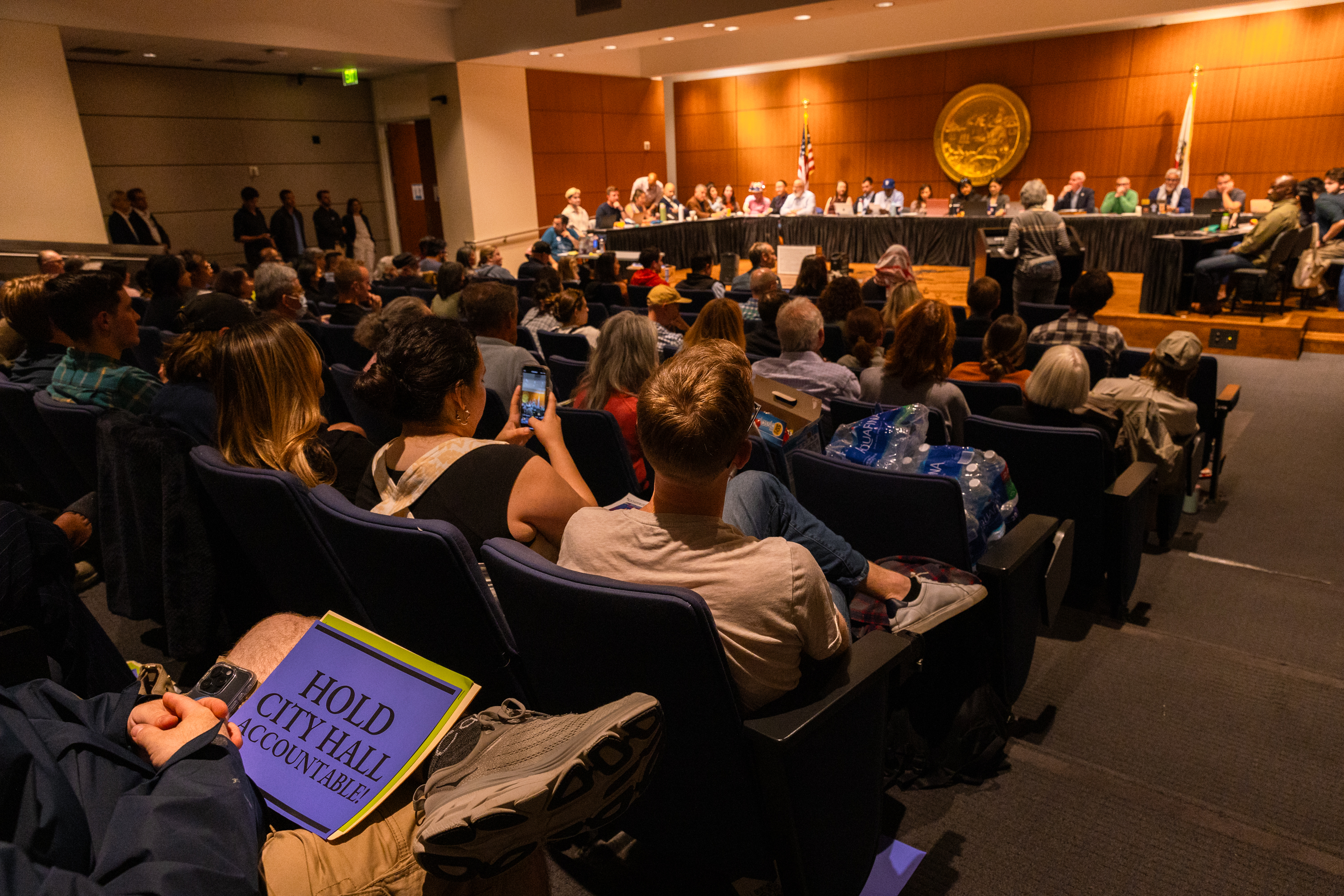 People sit in an auditorium, facing a long panel of officials. One person holds a sign reading “HOLD CITY HALL ACCOUNTABLE!” while others use their phones.