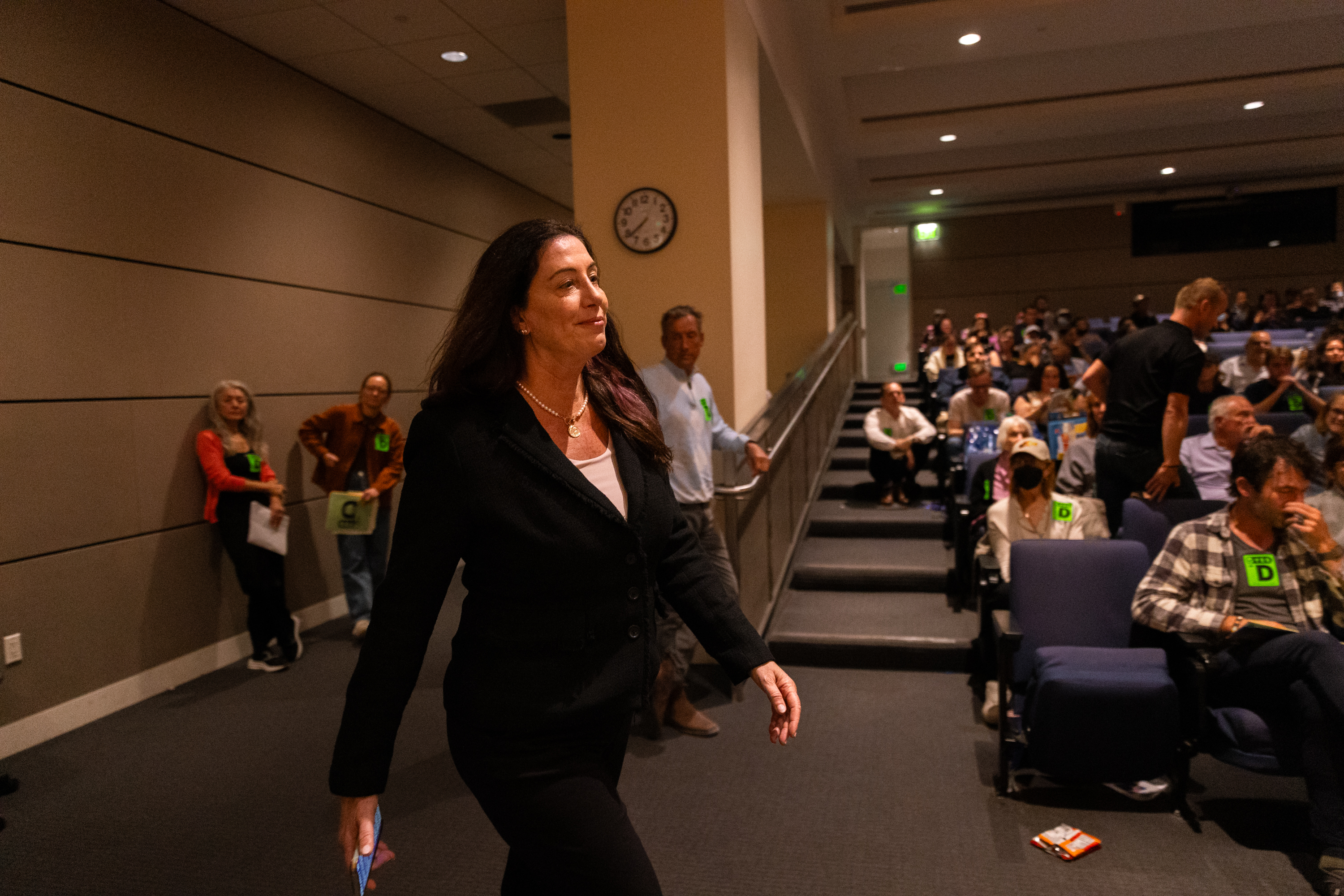 A woman in a black suit walks towards the front of a lecture hall, which has seated audience members and a clock on the wall. Other people stand nearby.