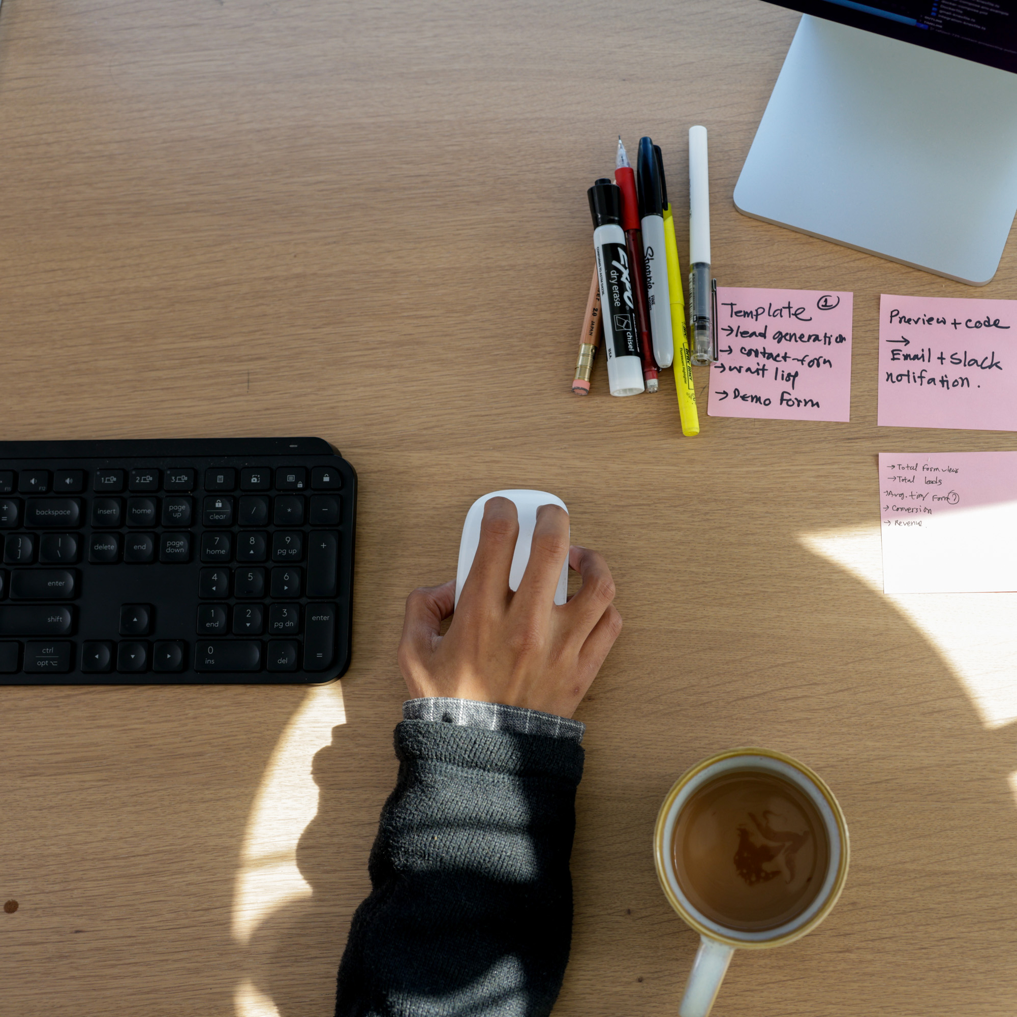 A hand using a computer mouse is shown next to a keyboard, sticky notes with tasks written on them, various pens, and a coffee cup on a wooden desk.