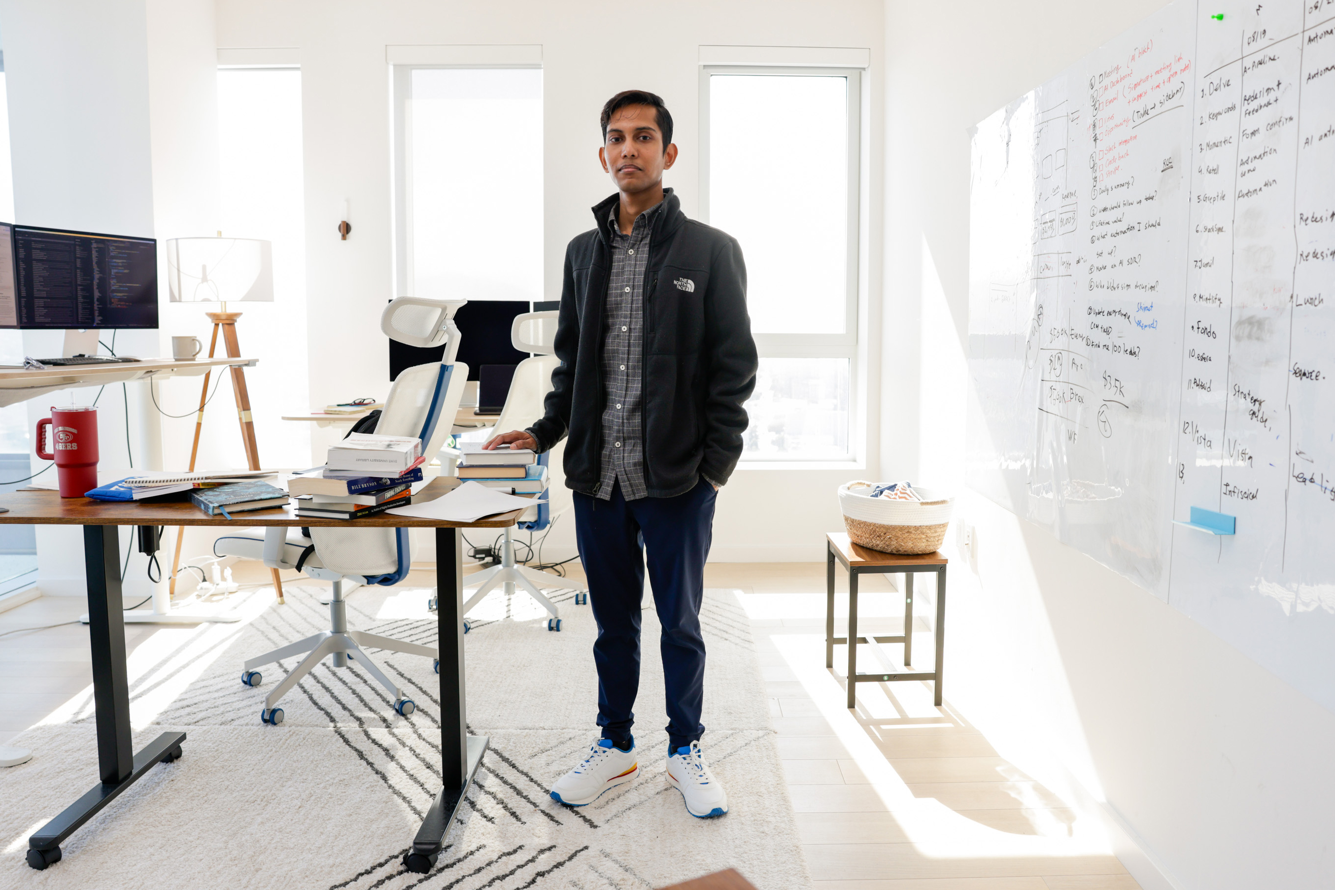 A man stands in a modern office with natural light. He is near a desk cluttered with books and a computer. A whiteboard with notes is on the right wall.