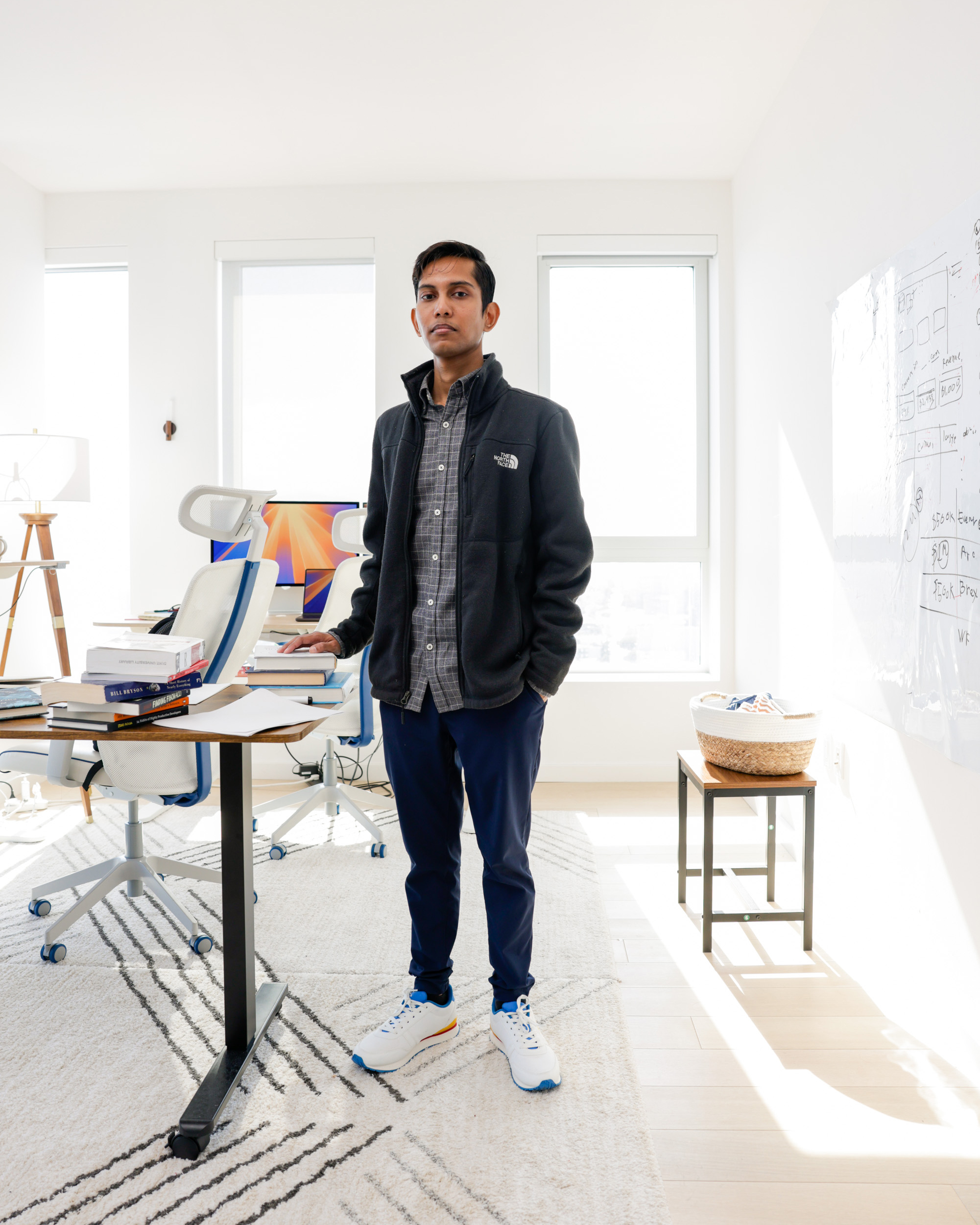 A man in smart-casual attire stands in a bright, modern office with a desk, books, computer, chairs, and a whiteboard on the wall.