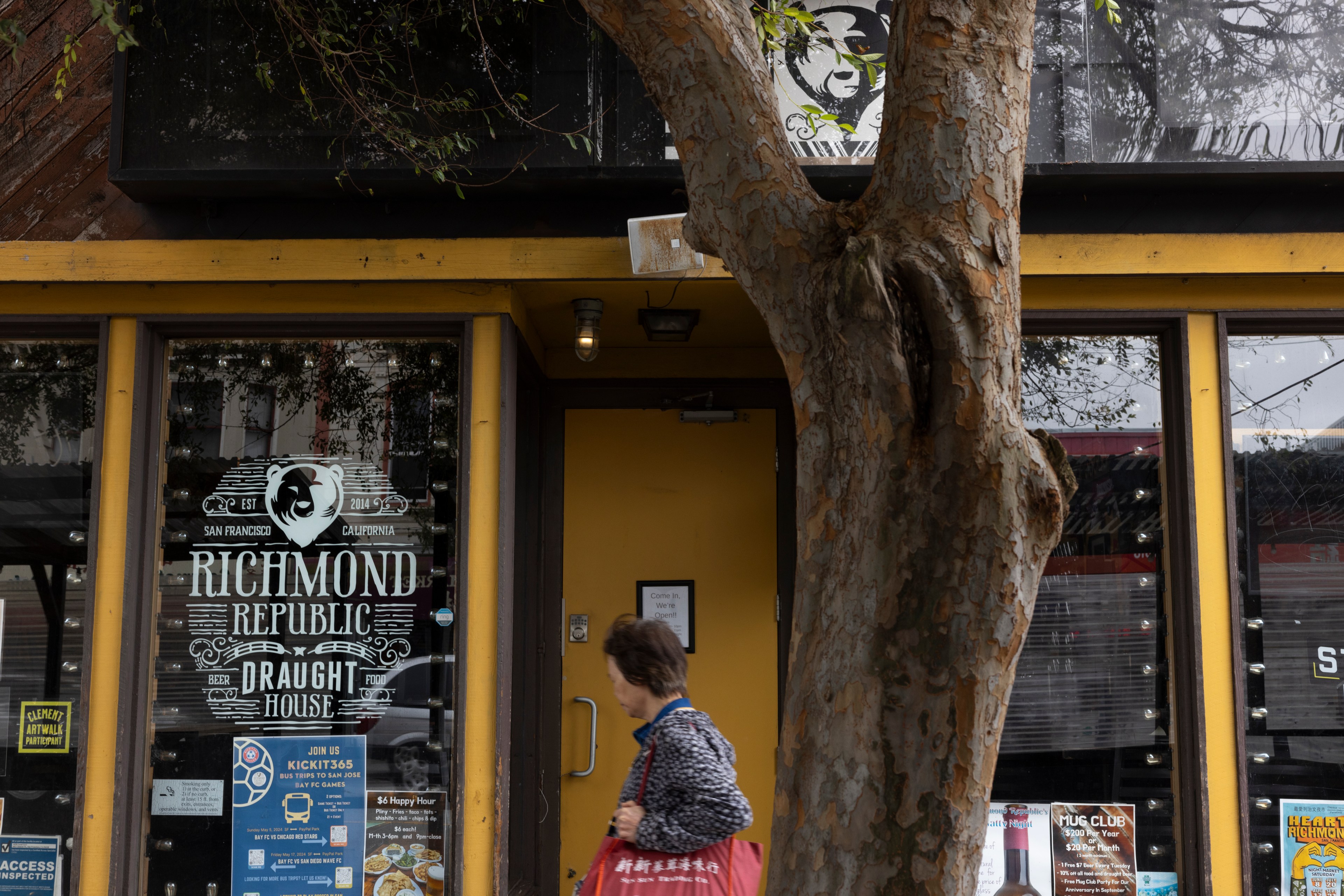 The image shows the entrance to "Richmond Republic Draught House" with glass doors, a large decal, and a yellow frame. A person with a red bag walks past a tree in the foreground.