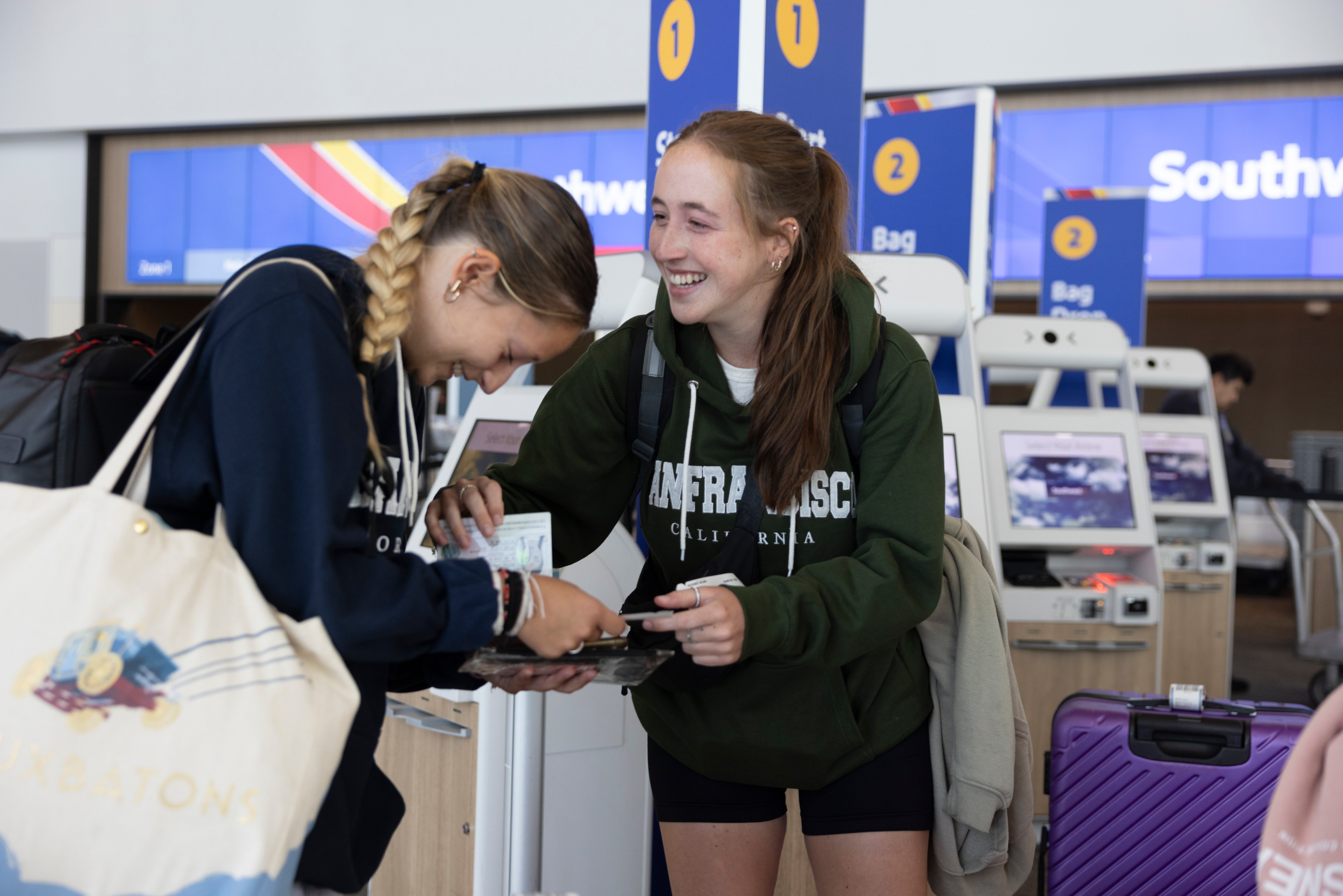 Two young women, smiling and laughing, check in at an airport kiosk with a large tote bag, backpack, and carry-on luggage. The sign behind them reads &quot;Southwest&quot;.