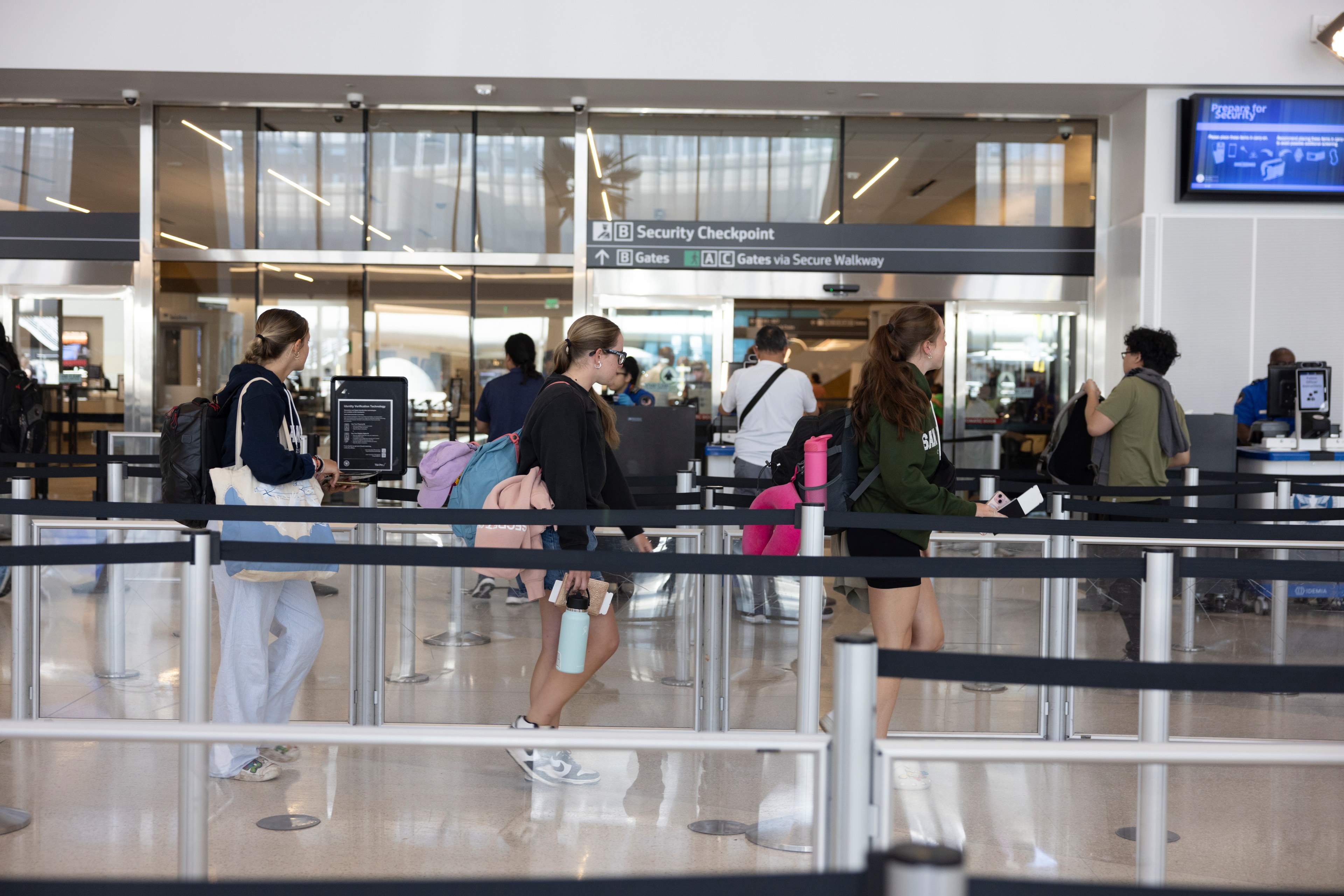 Passengers with baggage are waiting in a security checkpoint line at an airport, moving through a roped pathway towards the security screening area.